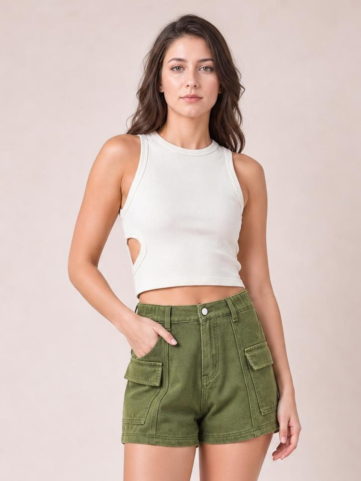 A woman modeling a sleeveless white crop top with a side cutout and olive green cargo shorts against a solid light beige background.