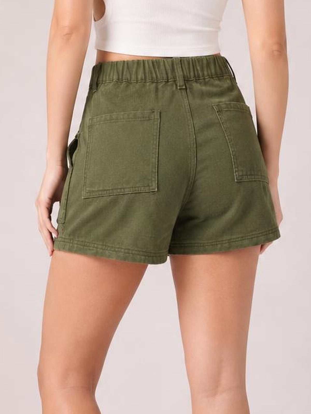 A model wears olive green high waist shorts with an elastic waistband, visible seam details, roomy rear patch pockets, and a relaxed mid-thigh silhouette.