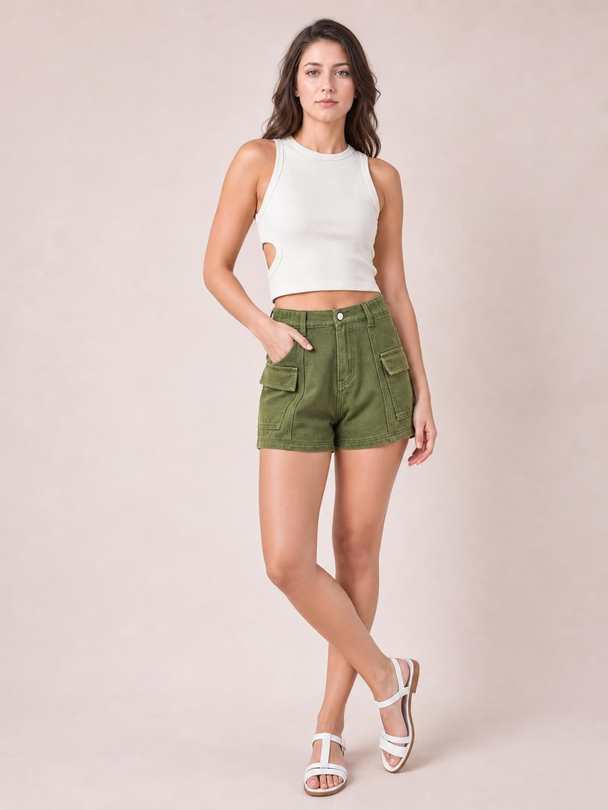 A model poses with crossed legs and one hand in olive green high-waist shorts featuring cargo pockets, belt loops, a button closure, and seam detailing.