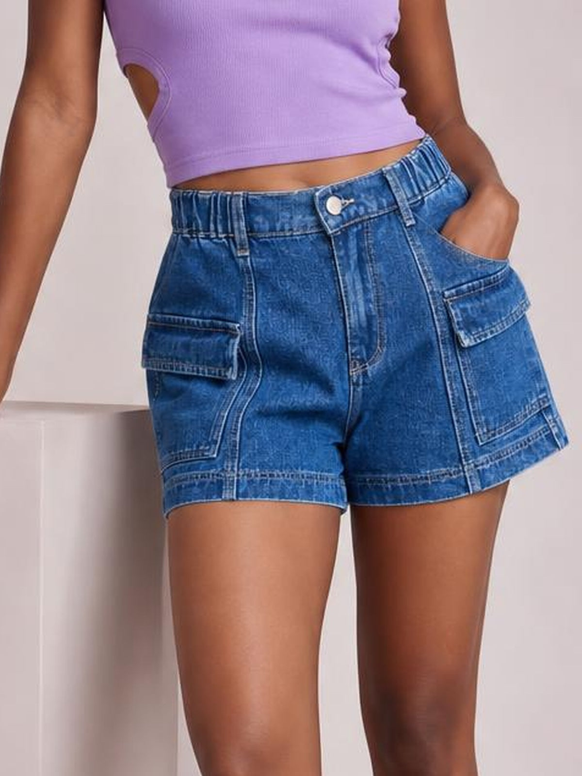 A standing model wears blue high-waist denim shorts with a relaxed silhouette, button fly, belt loops, contrast seams, and multiple utility pockets.