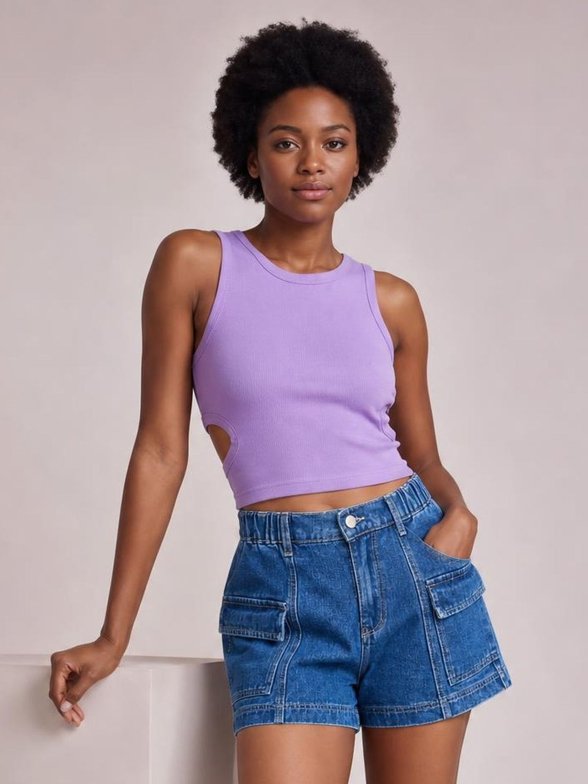 A woman modeling a lavender sleeveless crop top with side cutouts and blue denim shorts against a plain light beige studio background.