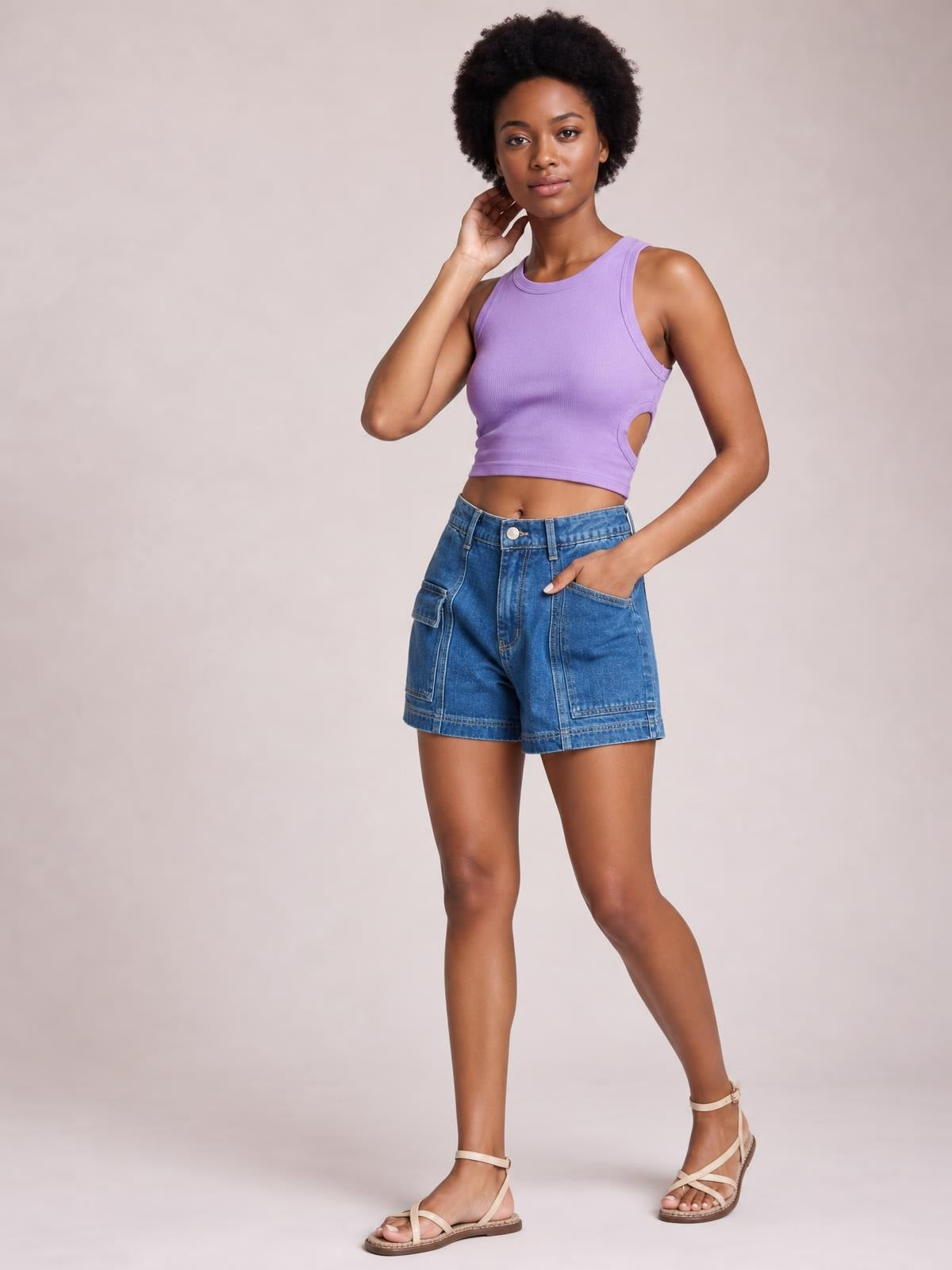 A model poses in blue high-waist denim shorts with seam details, a relaxed silhouette, and multiple patch pockets, one hand tucked into the side pocket.
