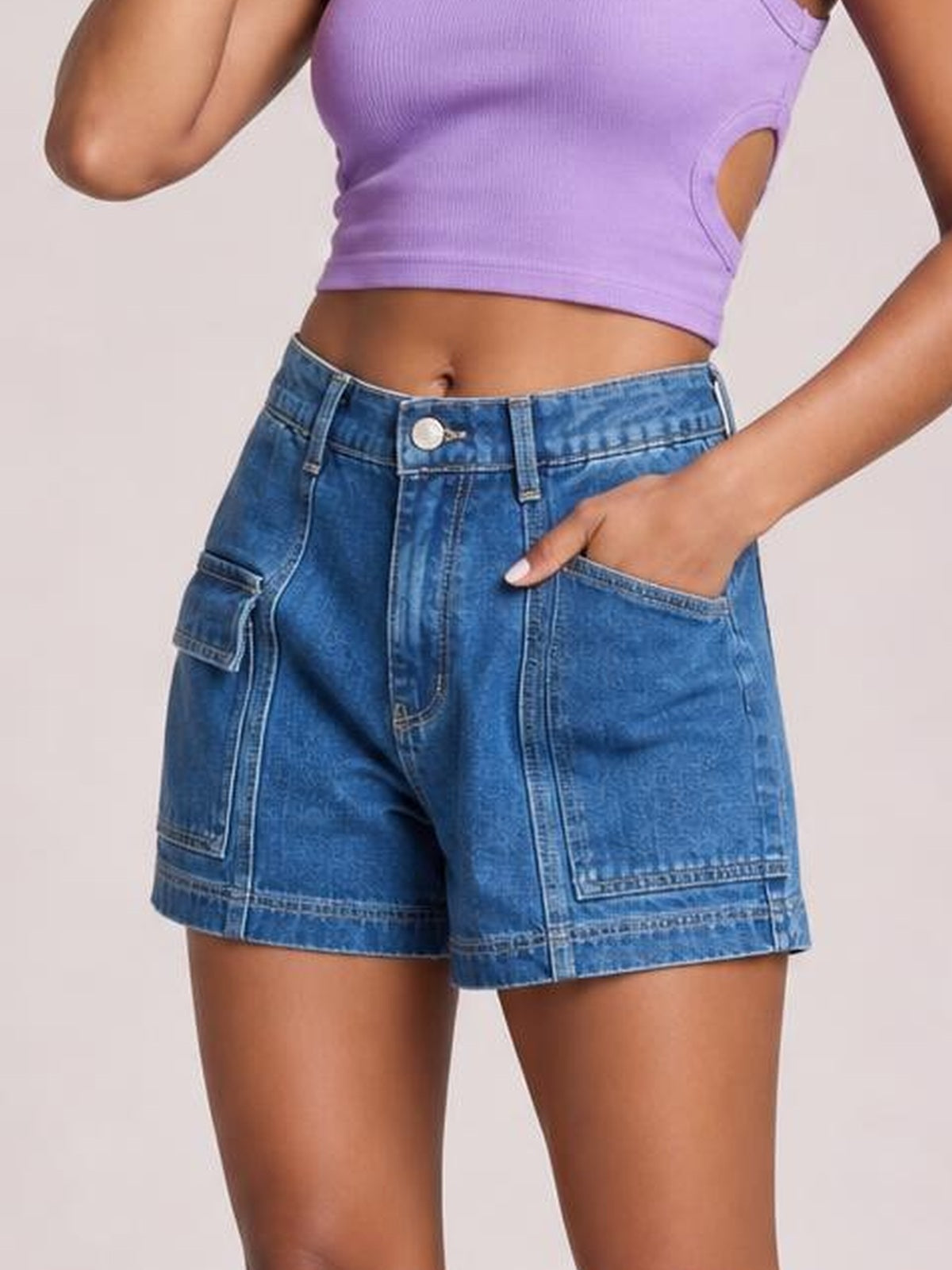 Model wearing blue high-waist denim shorts with a button fly, seam details, roomy straight silhouette, and multiple patch pockets, hand resting in one pocket.