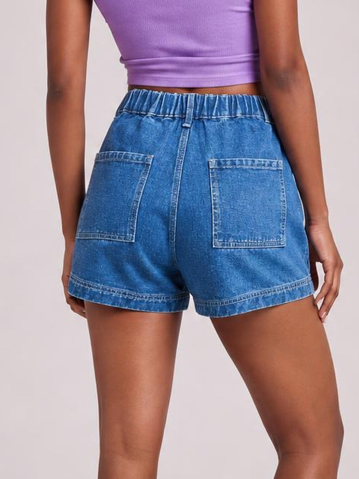 A model stands in blue high waist denim shorts with an elastic waistband, visible seam detailing, two large back patch pockets, and a relaxed leg silhouette.