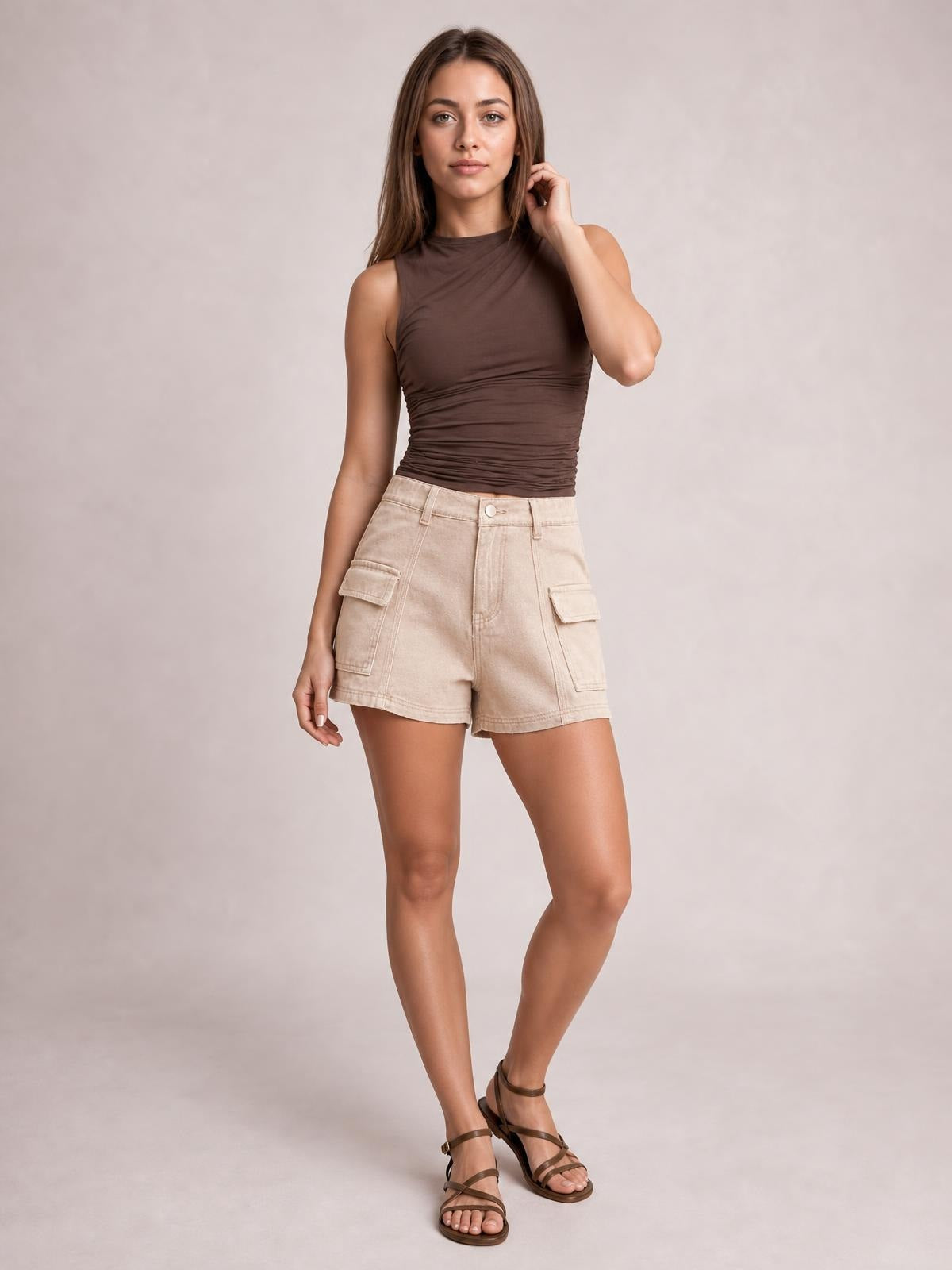 A model wears beige high-waist shorts with stitched seams, a button fly, and flap cargo pockets, standing with one leg crossed.