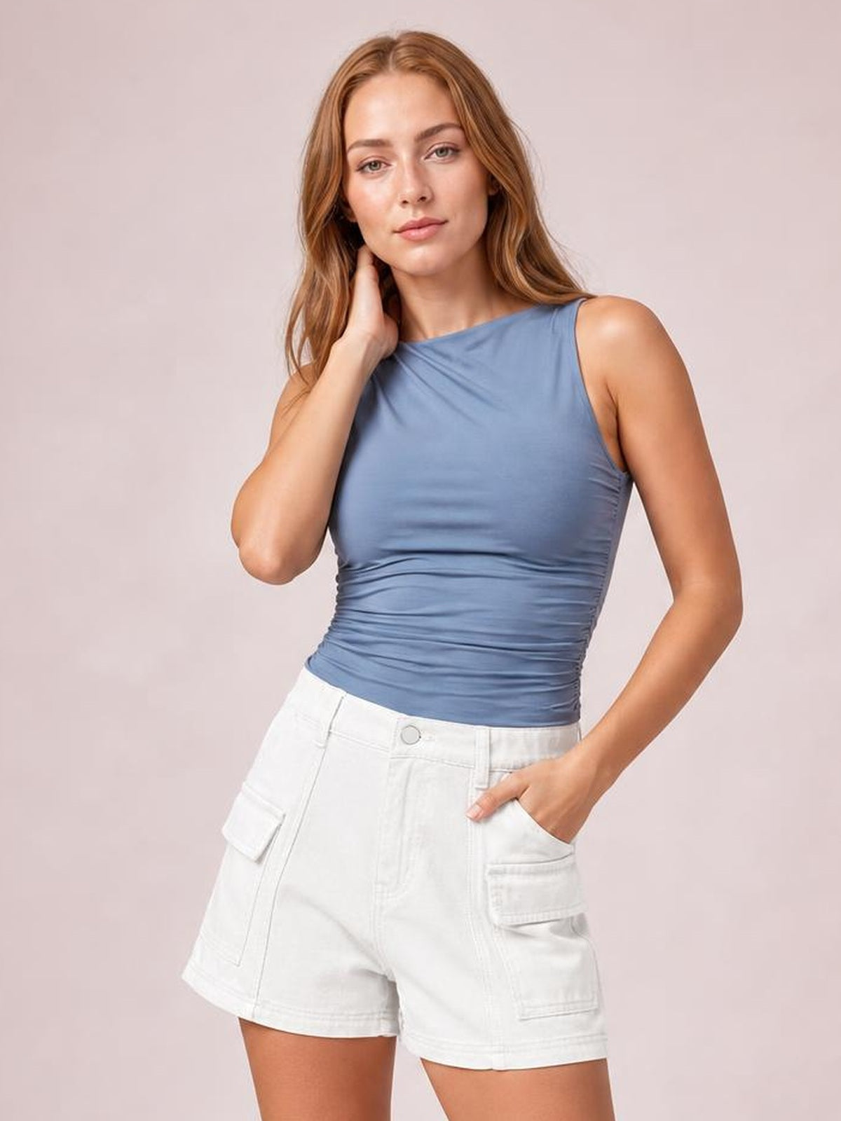 A woman modeling a sleeveless, ruched slate blue top paired with white cargo shorts against a solid light pink studio background.