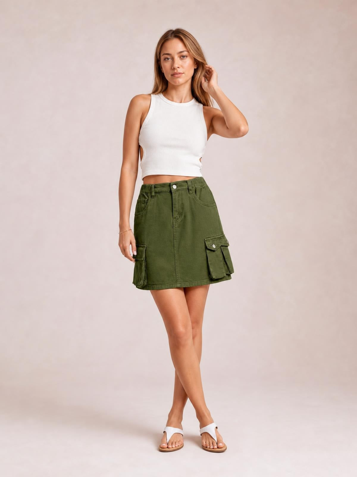 A model poses with crossed legs in an olive high-waist denim skirt featuring a straight mini silhouette, belt loops, front button closure, and cargo pockets.