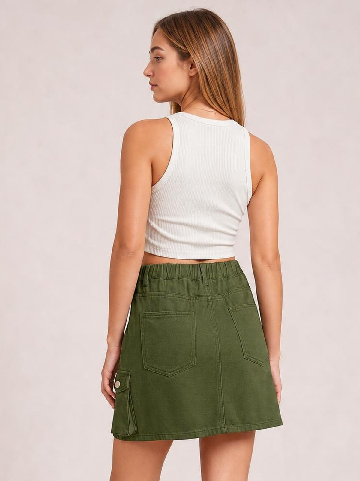 A model poses turned away in an olive green high-waist denim skirt with a straight silhouette, back patch pockets, and a buttoned cargo pocket.