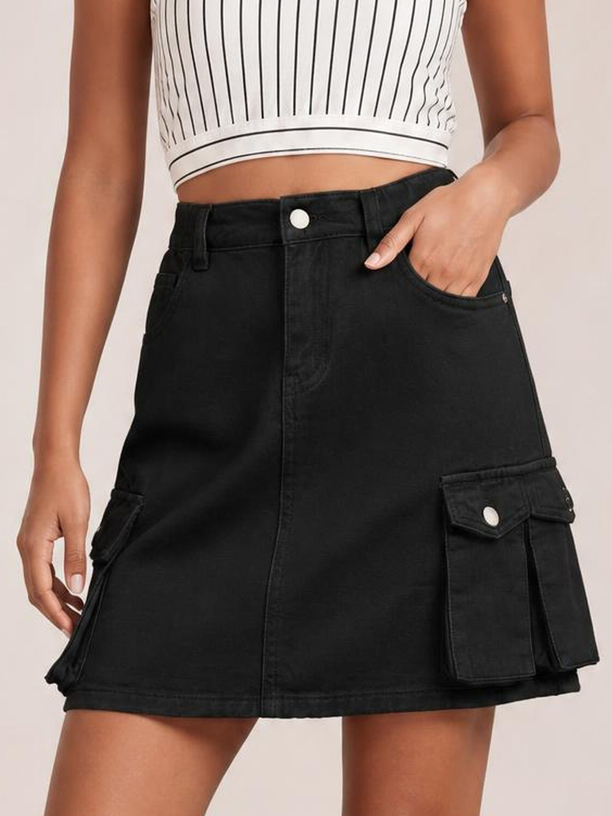 Model wears a black high-waist denim skirt with belt loops, front closure, straight silhouette, and roomy flap cargo pockets, posing with one hand in a pocket.