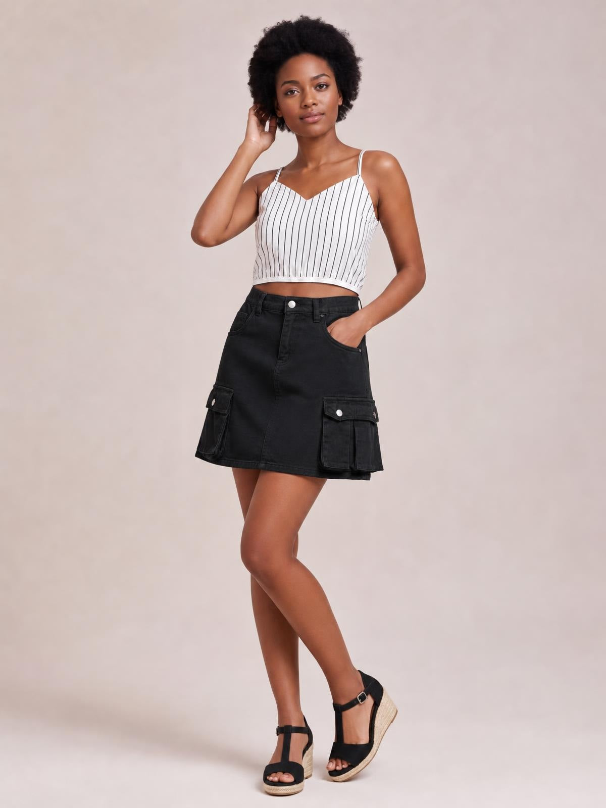 A model poses with one leg crossed in a black high-waisted denim mini skirt featuring front and cargo pockets, belt loops, and a straight silhouette.