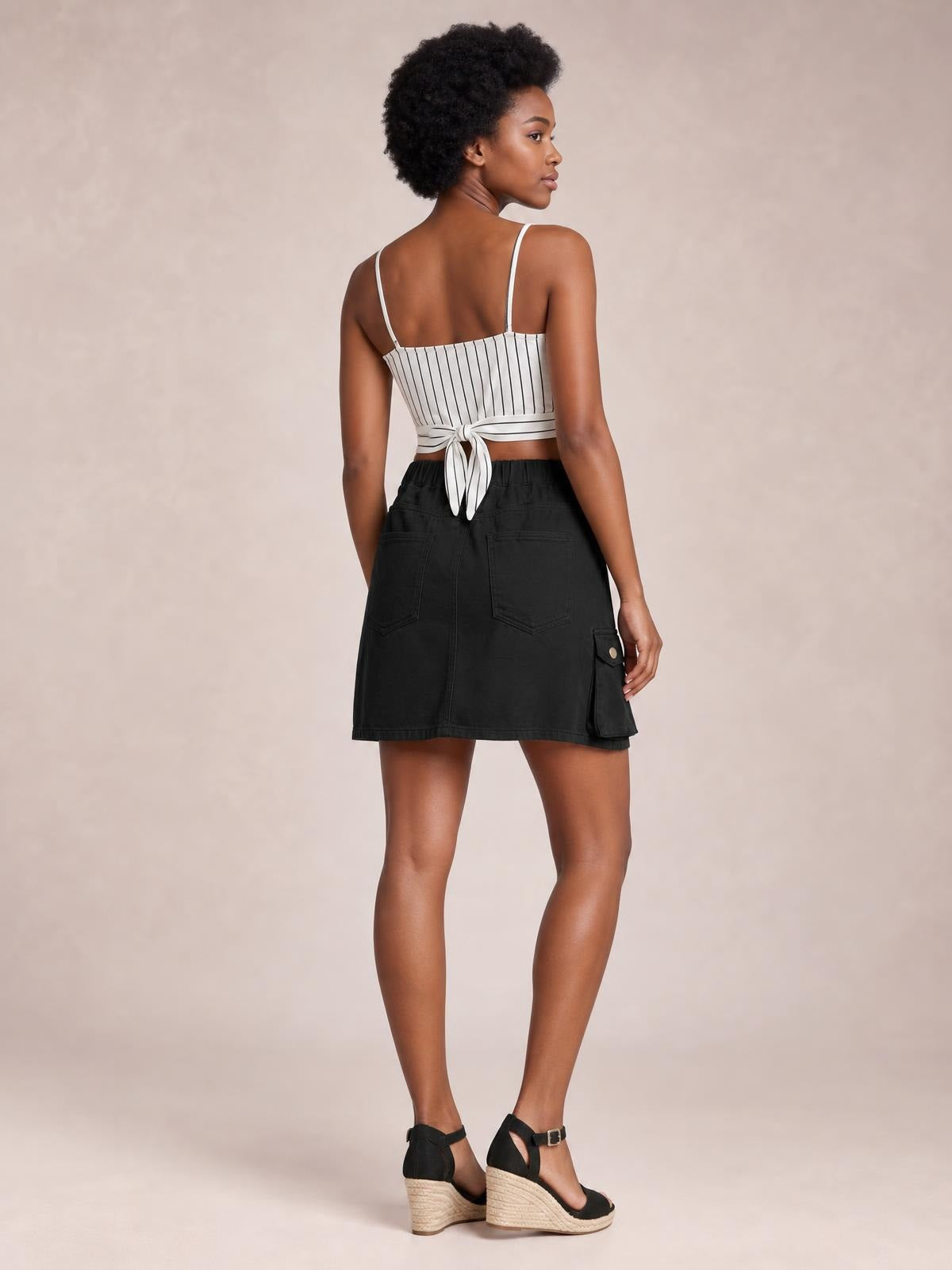A model looks over her shoulder wearing a black high waist denim skirt with a straight mini silhouette, back patch pockets, and a buttoned side cargo pocket.