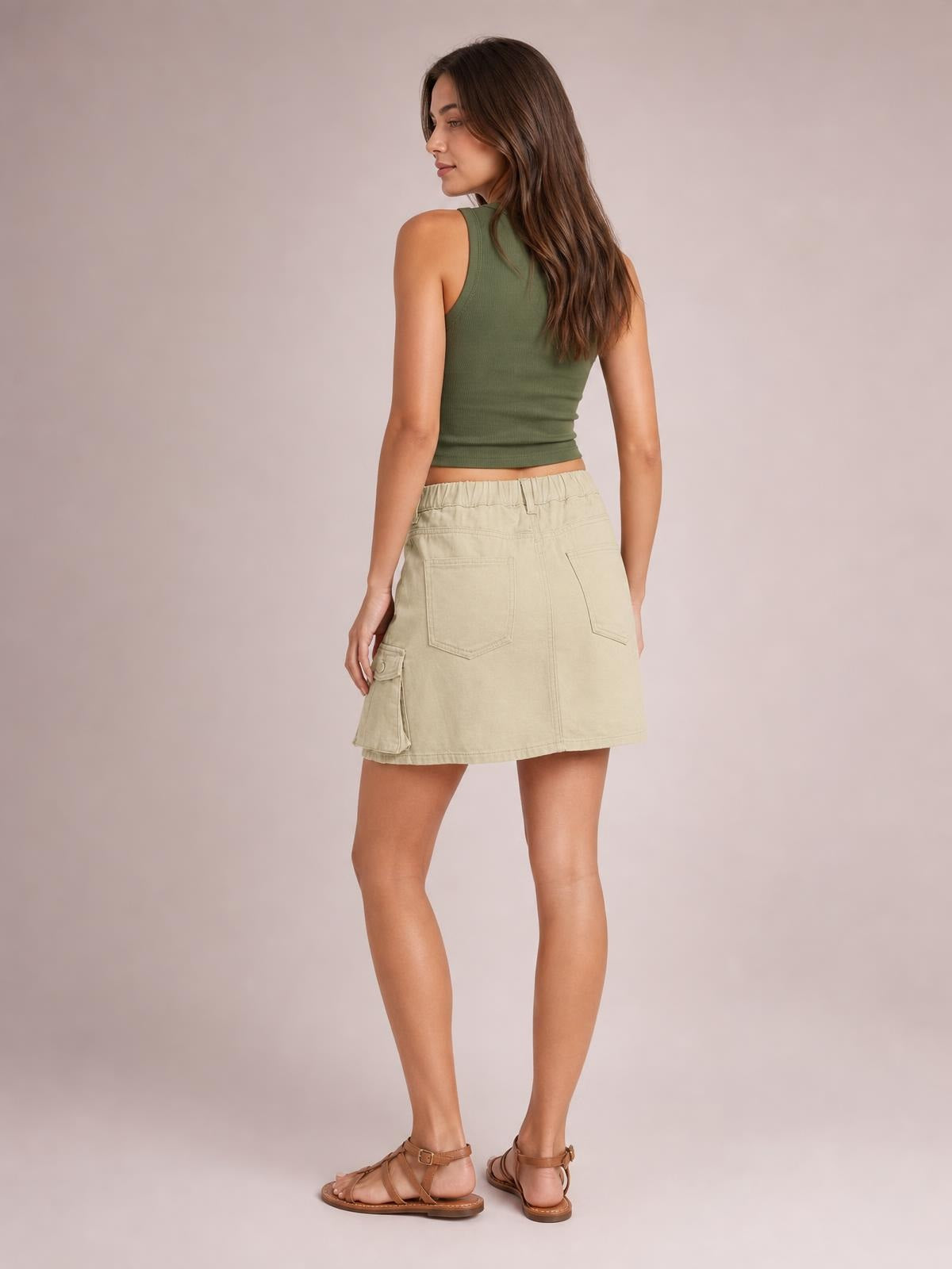 A model poses turned away in a high-waist beige denim skirt with rear patch pockets, a side cargo pocket, belt loops, and a straight mini silhouette.