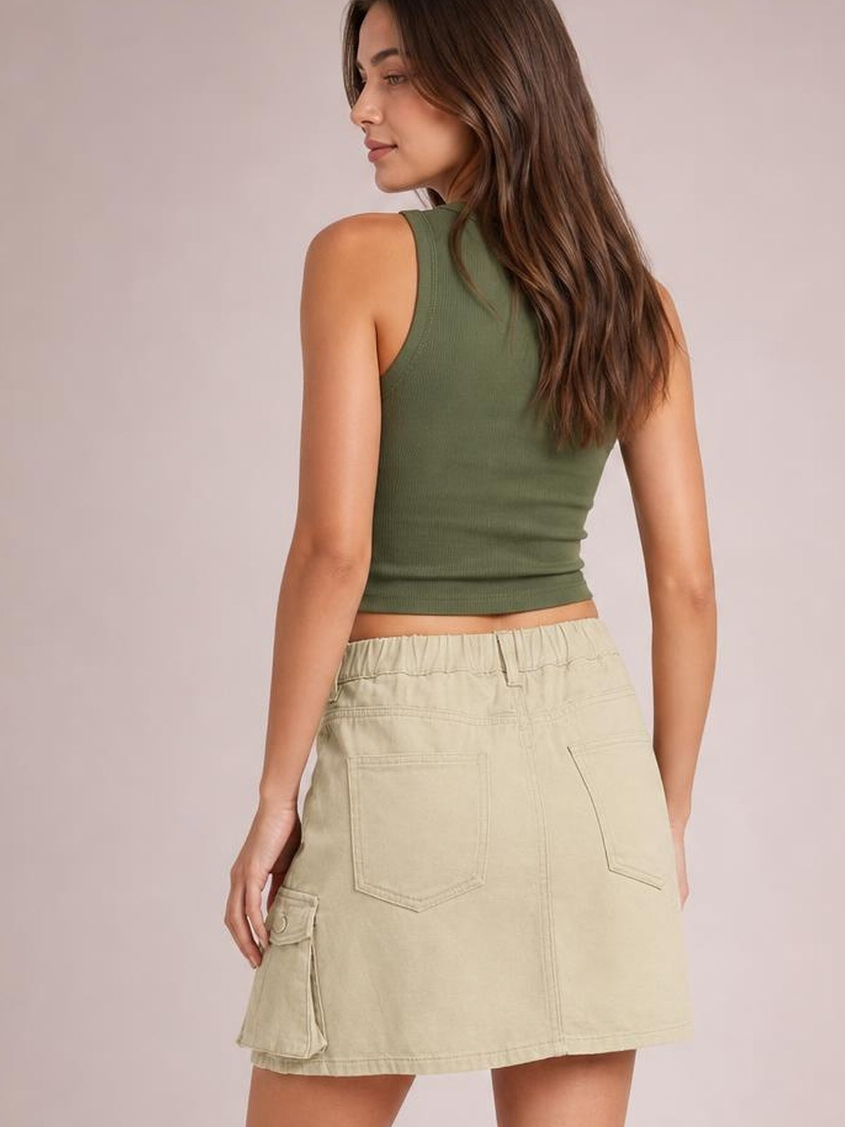 A model looks back over her shoulder in a beige high-waist denim skirt with a straight mini silhouette, back patch pockets, and a cargo side pocket.