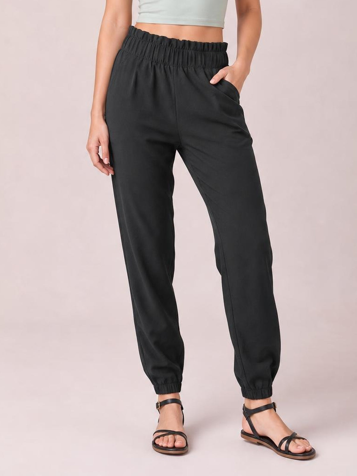 Model wearing black high waist joggers with an elastic waistband, side pockets, relaxed straight-leg fit, and elastic cuffs, standing with one hand in a pocket.