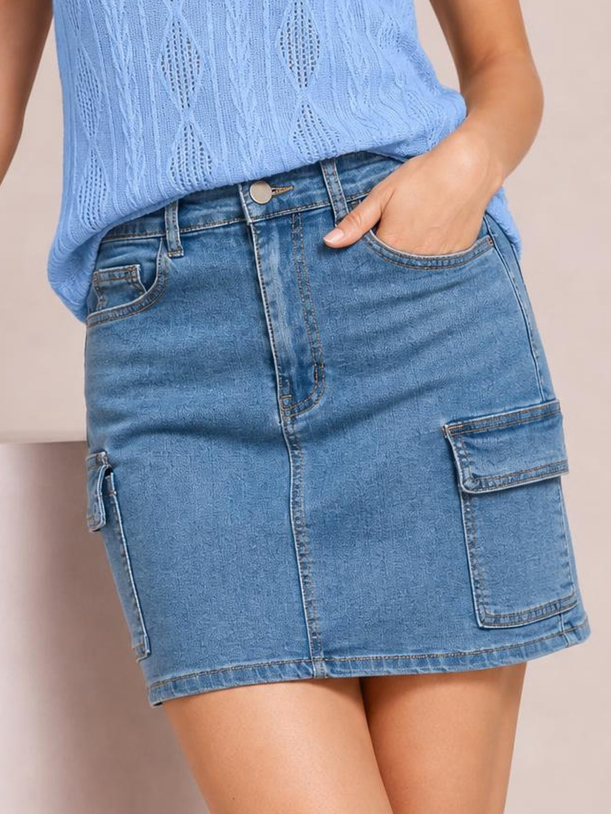 Model poses with one hand in the pocket of a high-waist blue denim skirt featuring belt loops, front pockets, cargo patch pockets, contrast stitching, and a straight mini silhouette.