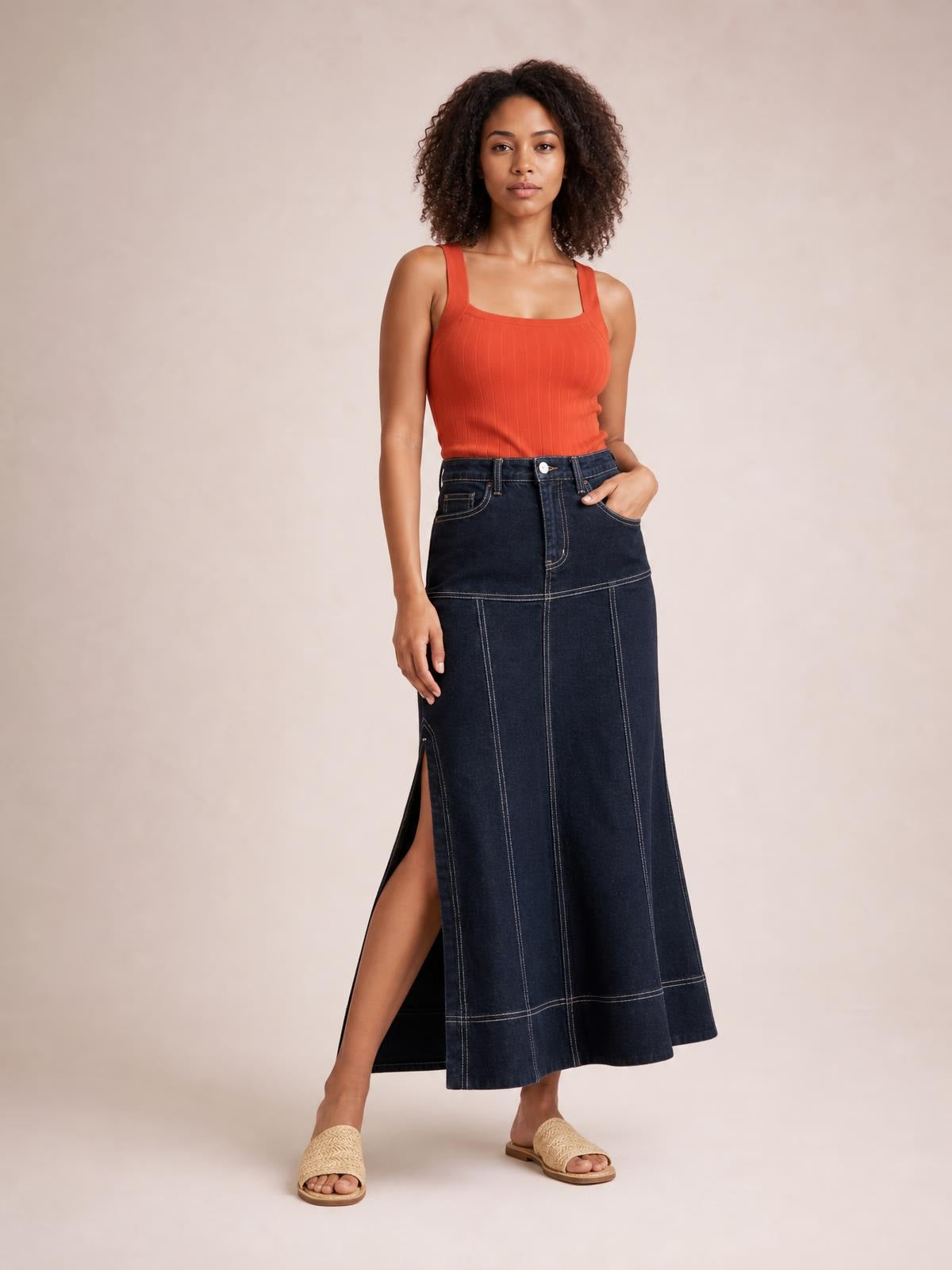 Model stands with one leg extended, wearing a dark denim high-waist maxi skirt with contrast stitching, exposed seams, and a dramatic side slit.
