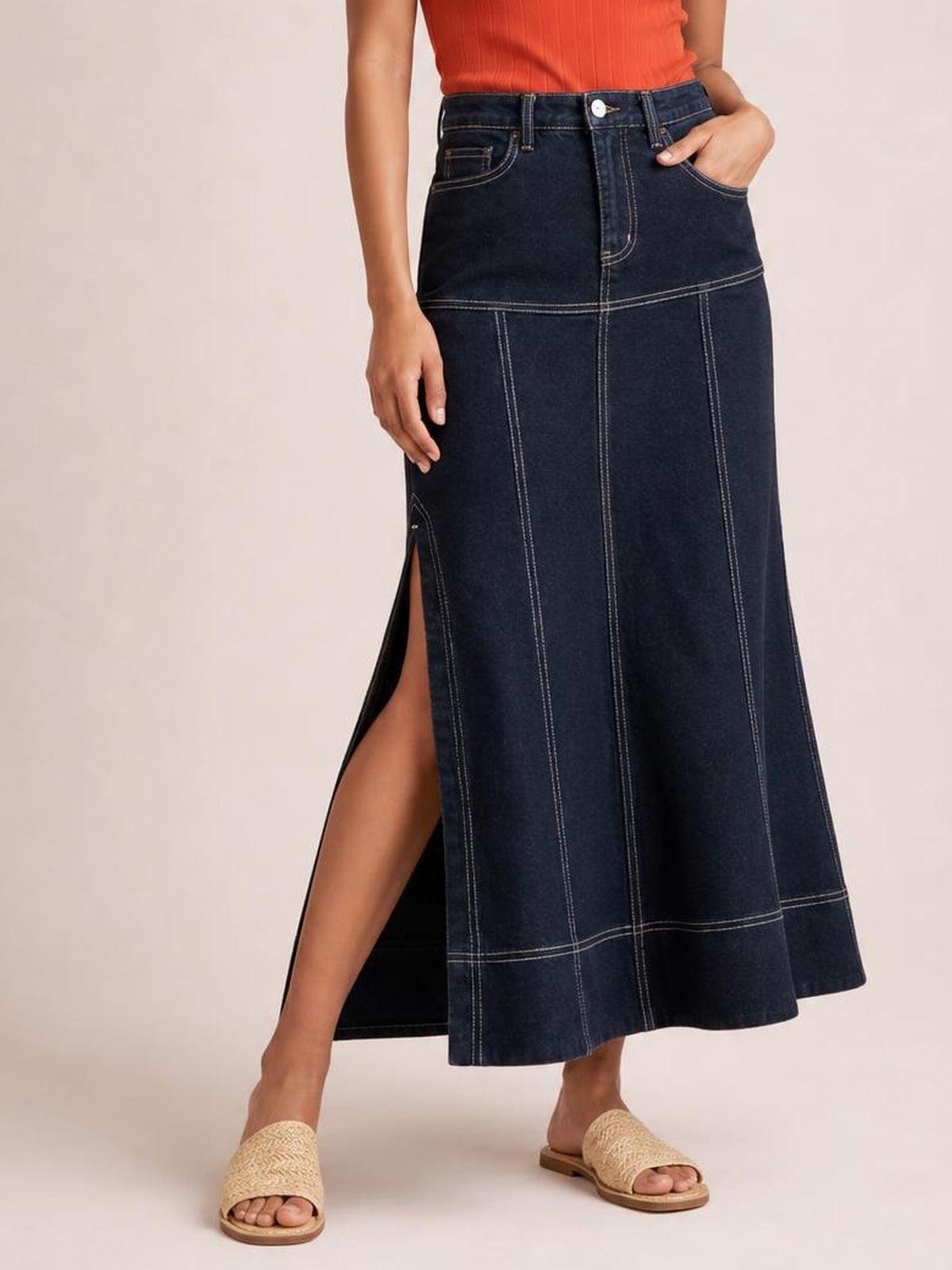Model wearing a high-waist dark denim maxi skirt with contrast exposed stitching, vertical seam panels, front pockets, and a side slit, standing with one hand in a pocket.