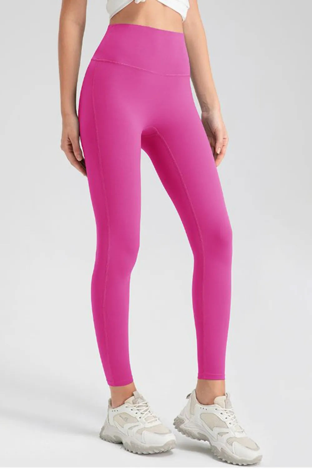 Model standing with hands at her sides wears bright pink high-waist skinny active pants with a wide waistband, seamed construction, and ankle-length silhouette.