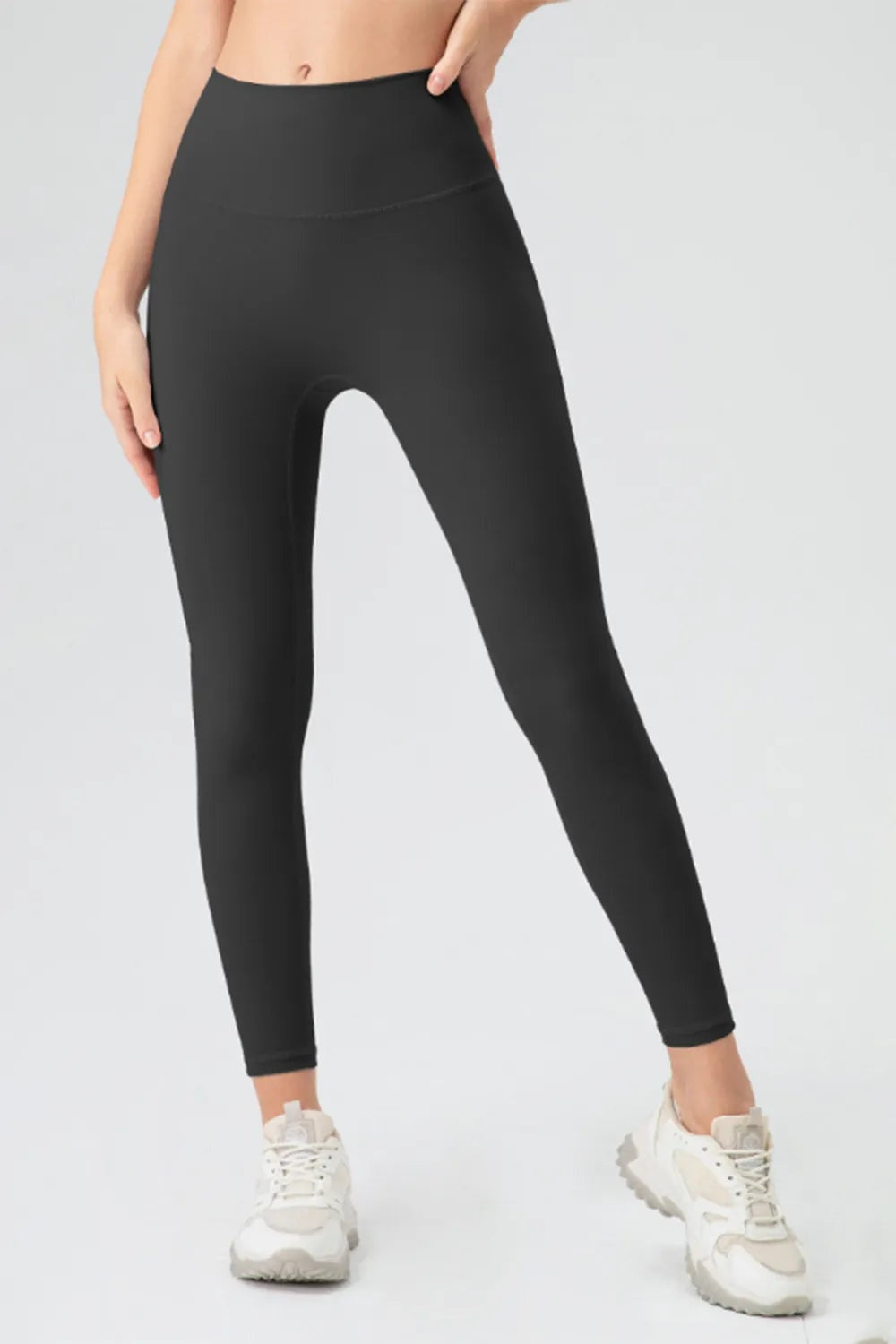 Model wears black high-waist skinny active pants with a smooth, fitted ankle-length silhouette and curved waistband seam, standing with one leg angled outward.