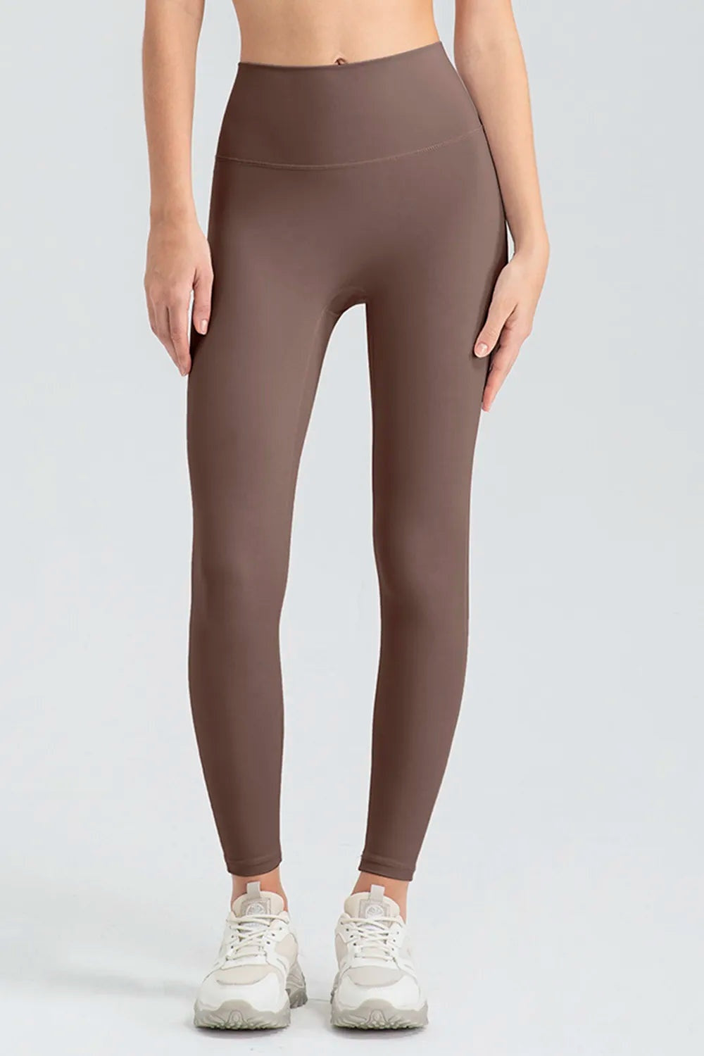 Model standing in brown high-waist skinny active pants with a smooth waistband, fitted full-length silhouette, and visible curved seam across the hips.