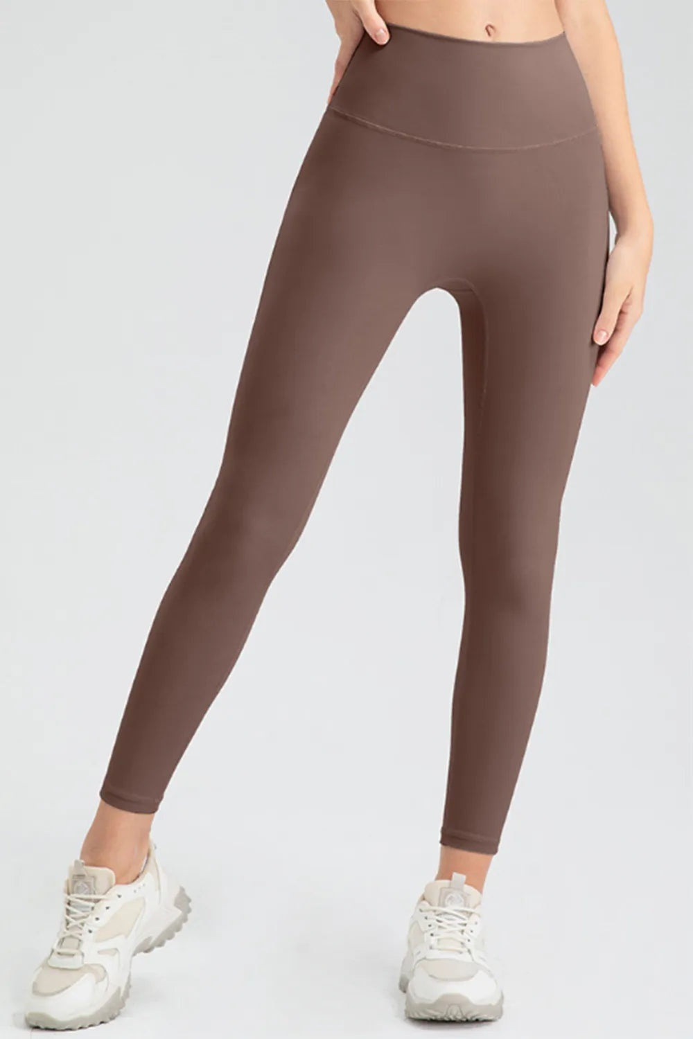 A model stands with legs apart wearing brown high-waist skinny active pants with a smooth wide waistband, curved hip seam, and fitted ankle-length silhouette.