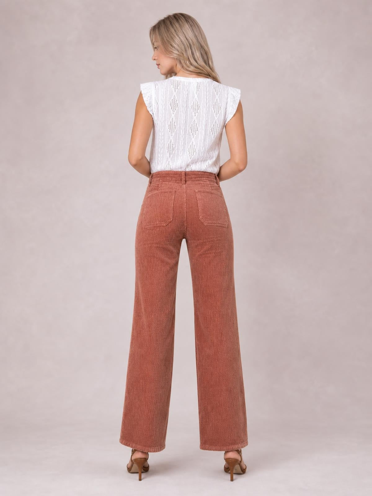 A model stands facing away in high-waist rust corduroy pants with square back pockets, belt loops, and a relaxed wide-leg silhouette.