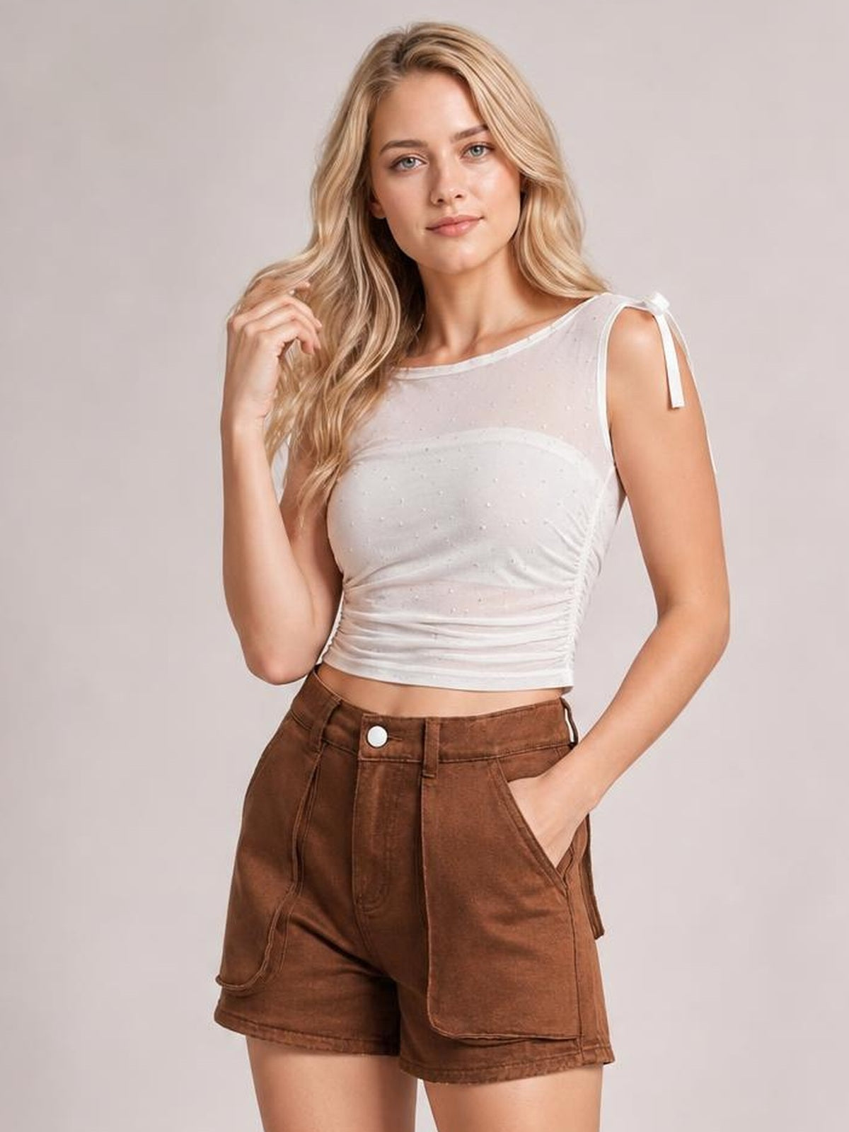 A blonde woman modeling a white sheer mesh crop top with side tie details and brown corduroy shorts against a light gray studio background.