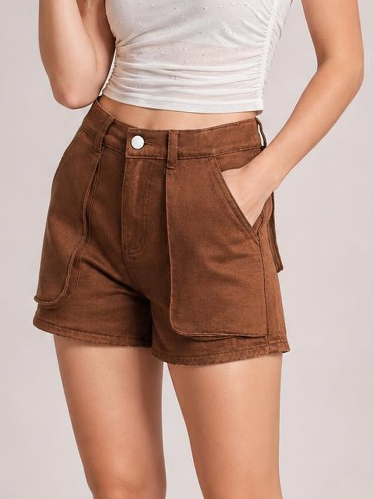 A model poses in brown high-waist utility denim shorts with a V-front waistband, button closure, belt loops, and oversized patch pockets, one hand tucked inside.