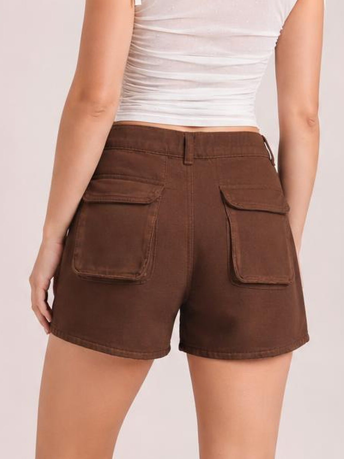 Back view of a model wearing brown high-waist utility pocket denim shorts with belt loops, large flap cargo pockets, and a relaxed Bermuda-length silhouette.