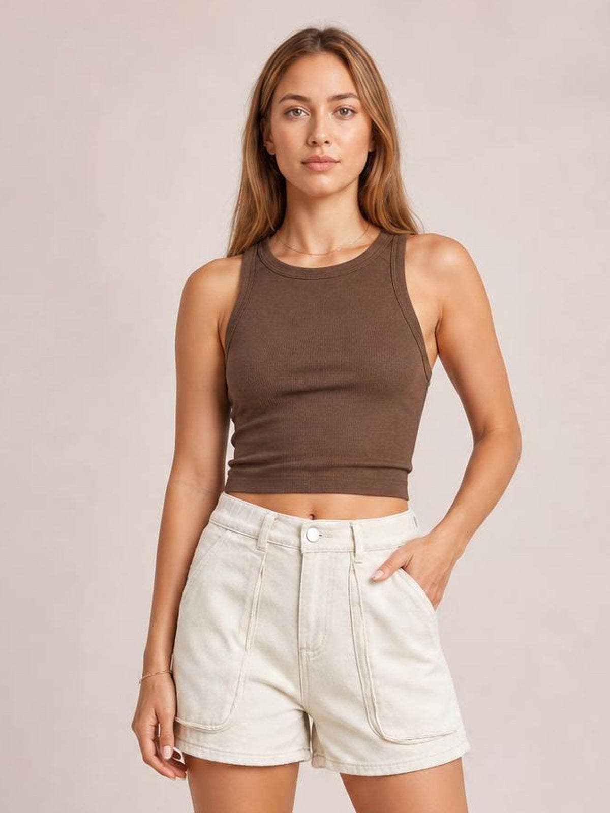 A woman modeling a brown ribbed sleeveless crop top paired with off-white denim shorts, set against a solid light beige studio background.
