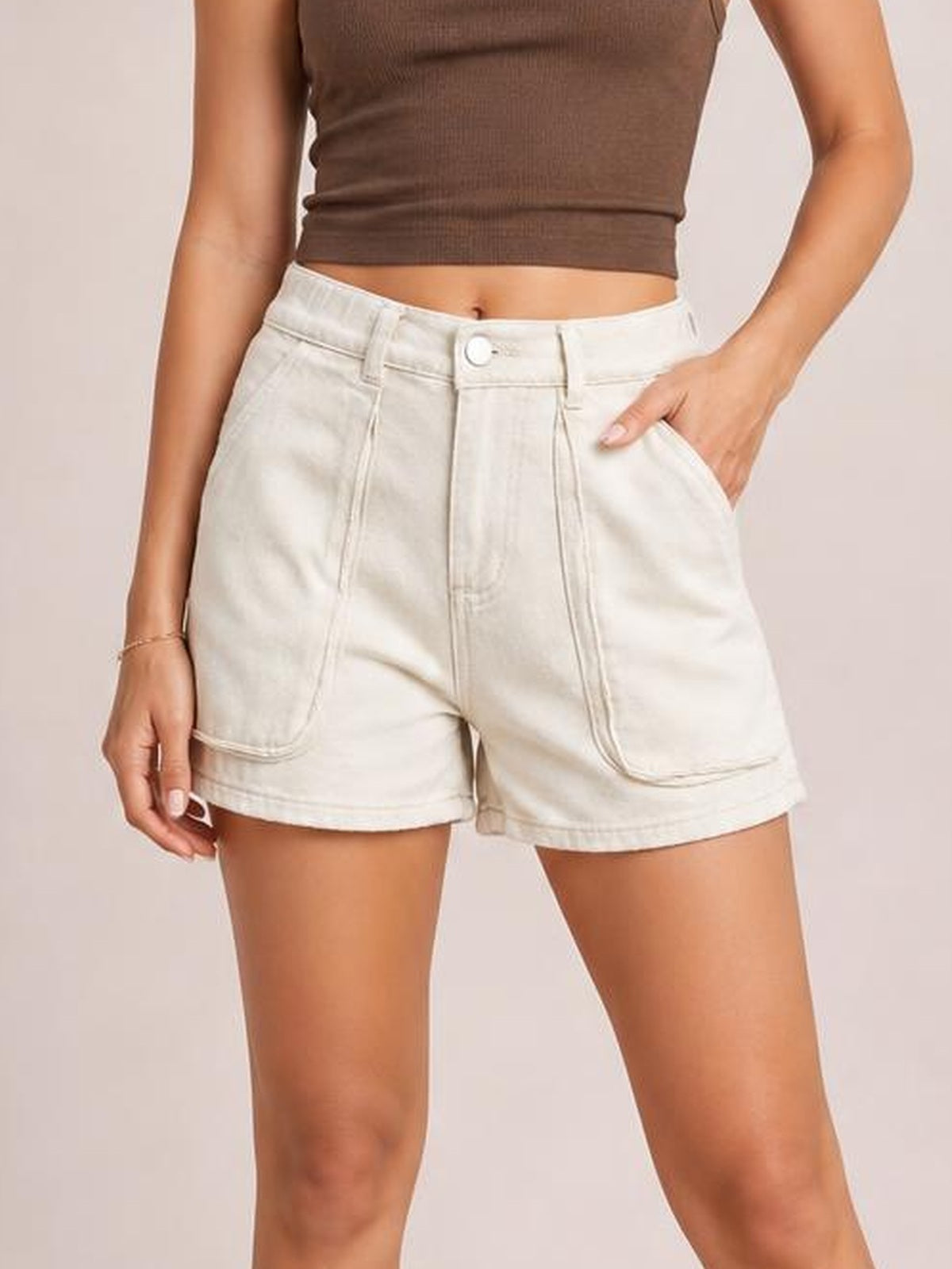 Model wearing high-waist ivory denim shorts with a V-front waistband, belt loops, button closure, roomy utility patch pockets, and a relaxed straight-leg silhouette.