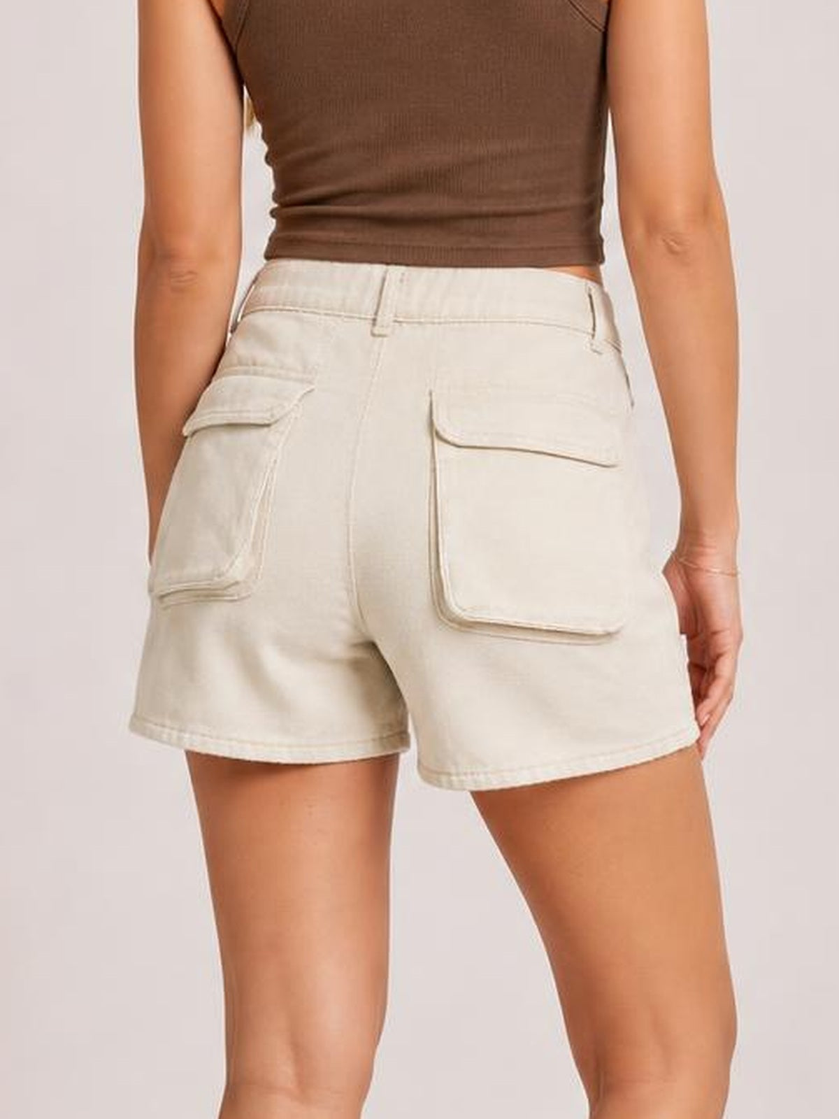 Back view of a model wearing cream high-waist utility denim shorts with belt loops, flap cargo pockets, stitched construction, and a relaxed Bermuda silhouette.