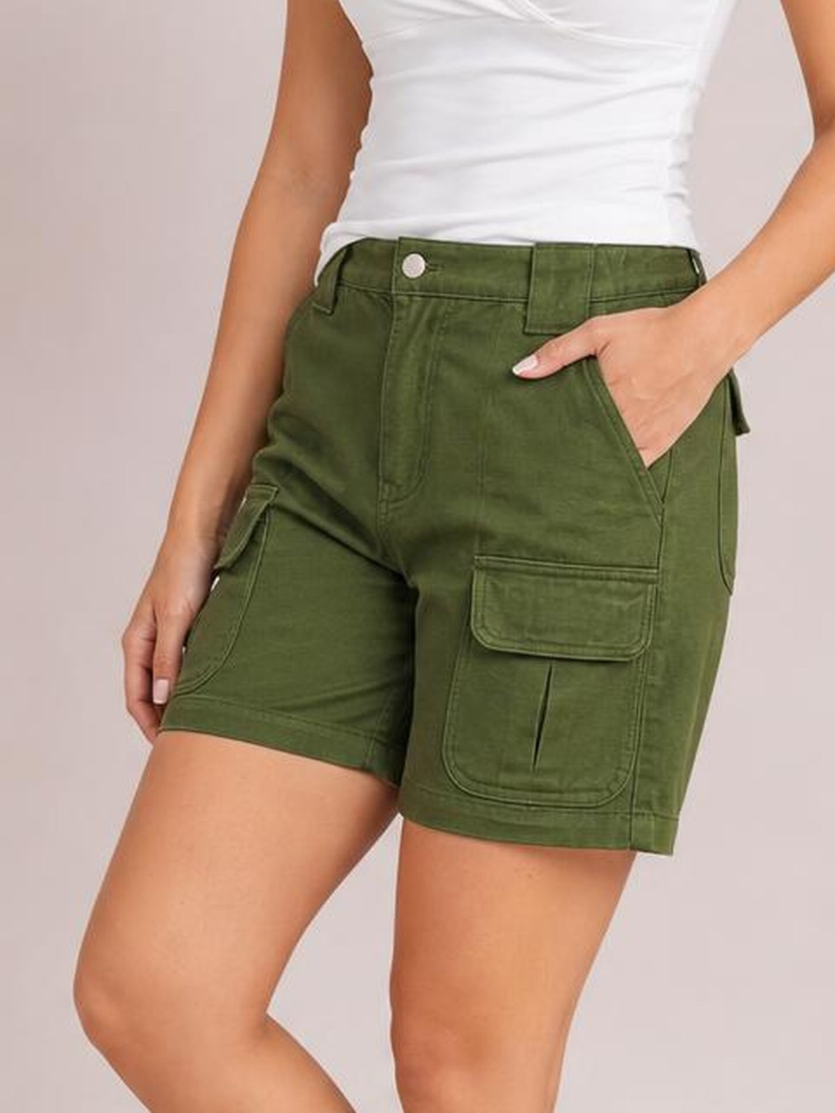 A model wears olive green high-waist washed denim shorts with belt loops, a button closure, utility pockets, and a straight-leg silhouette, posing with one hand in a pocket.