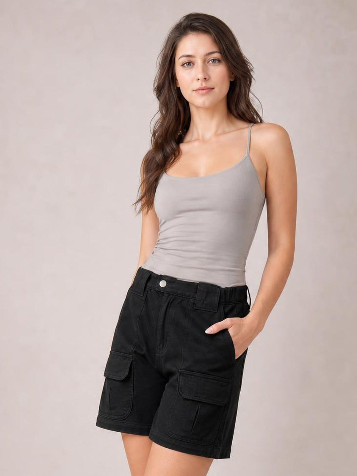 A woman modeling a light gray spaghetti-strap camisole top and black cargo shorts against a neutral beige studio background.