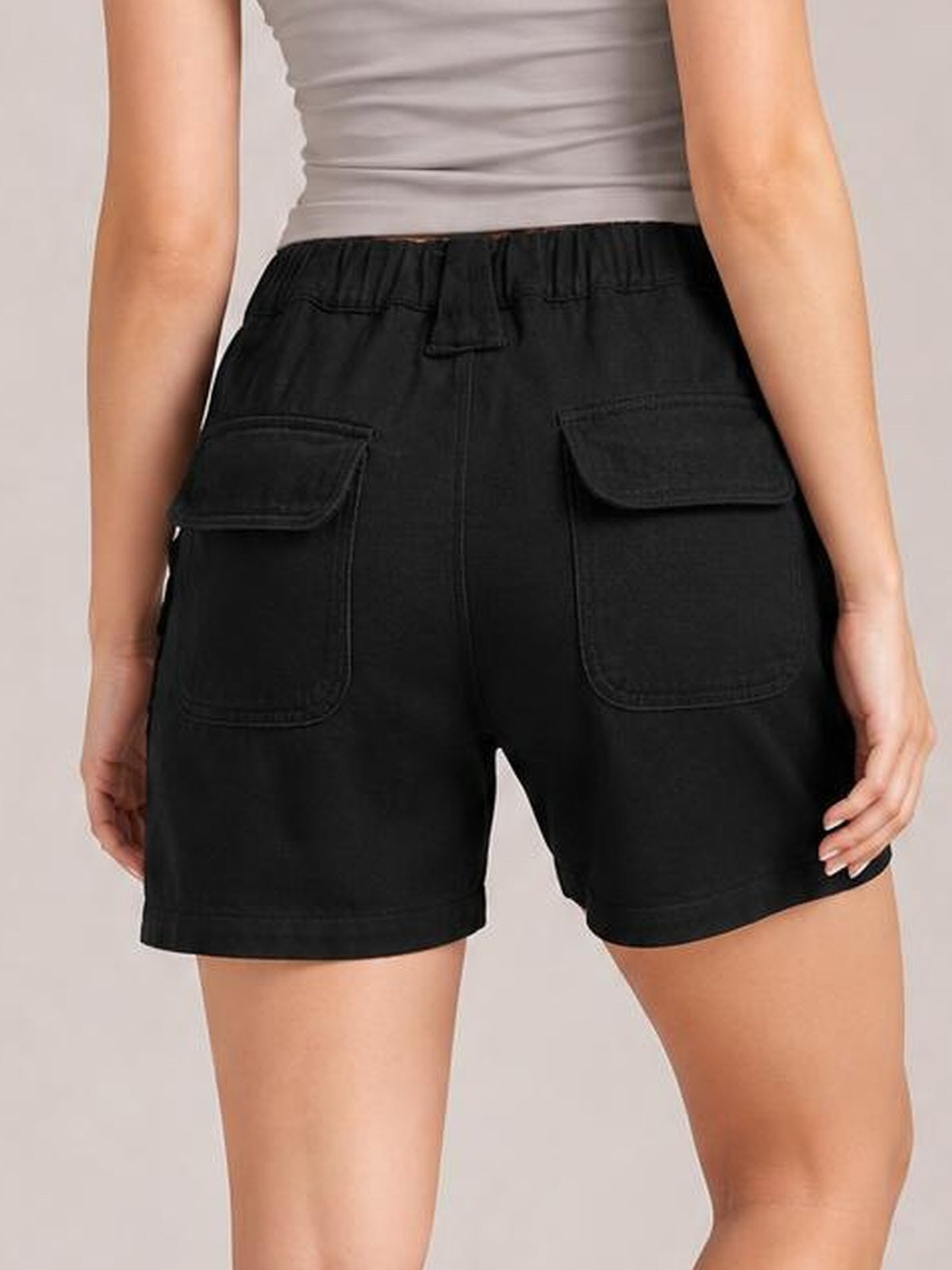 A model shown from behind wears black high-waist shorts with an elastic waistband, relaxed silhouette, stitched seams, and large back patch pockets.
