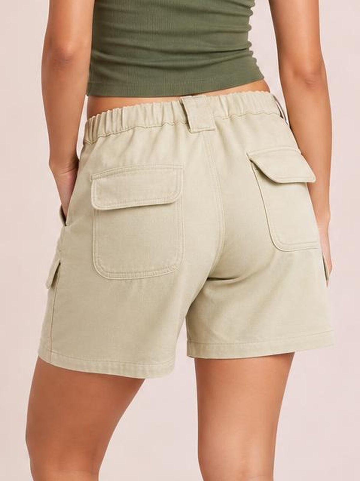 A model wears light beige high-waist washed denim shorts with an elastic waistband, straight silhouette, stitched seams, and multiple flap cargo pockets.