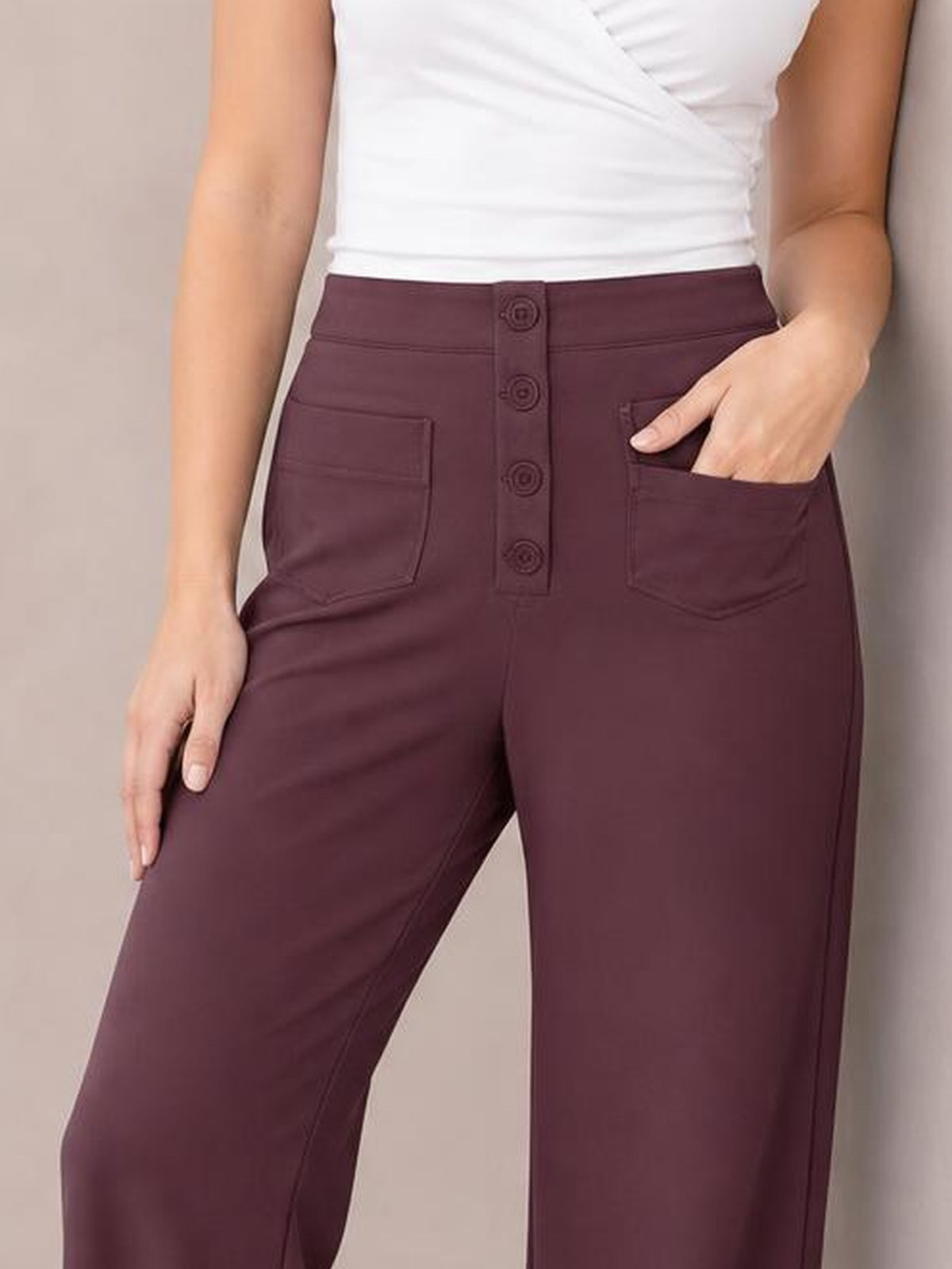 Model wears burgundy high waist wide leg pants with a button-front closure and patch pockets, posing with one hand tucked into a pocket.