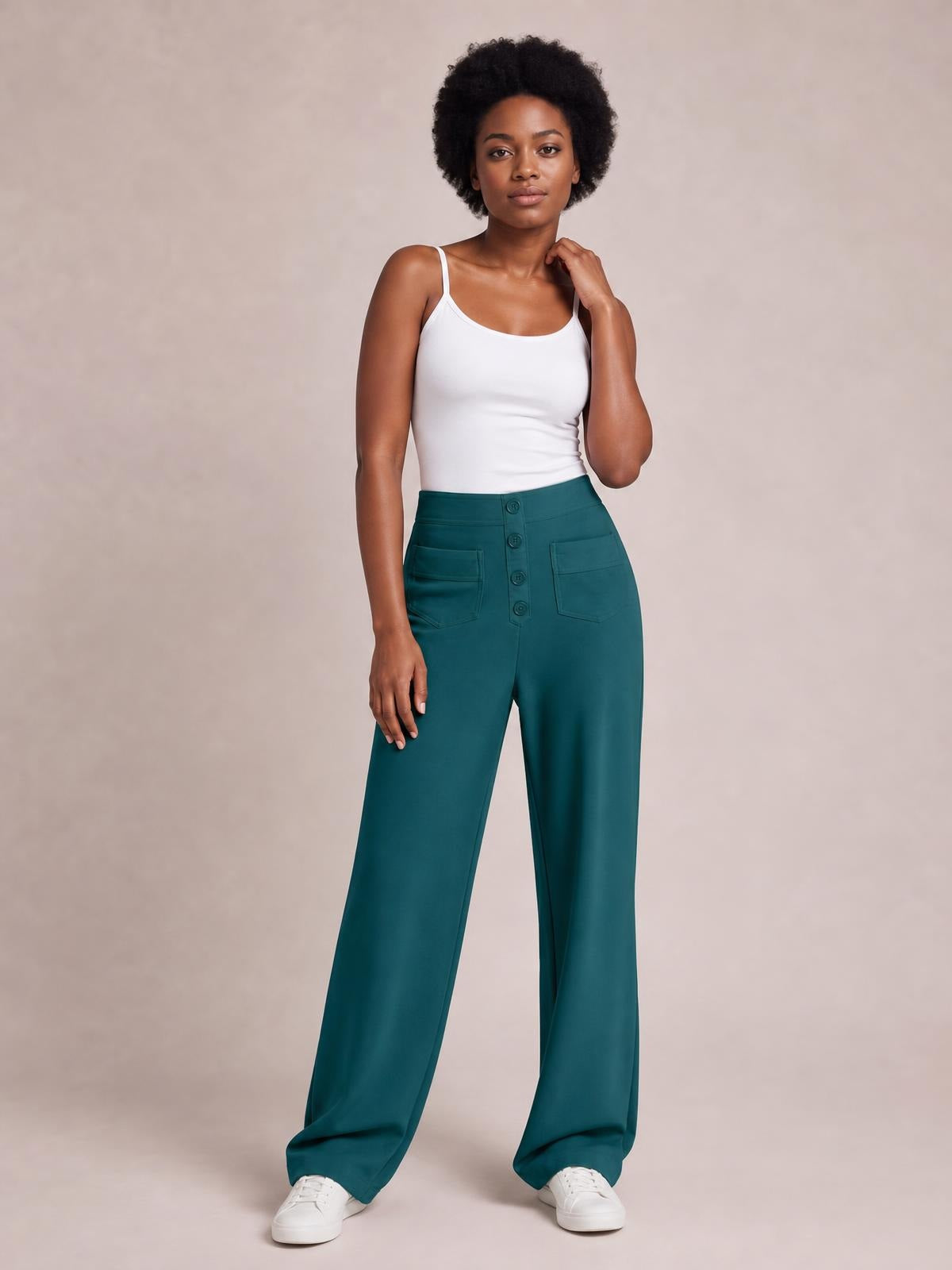 Model wears teal high waist wide leg pants with front button closure and patch pockets, standing with one hand raised near her shoulder.