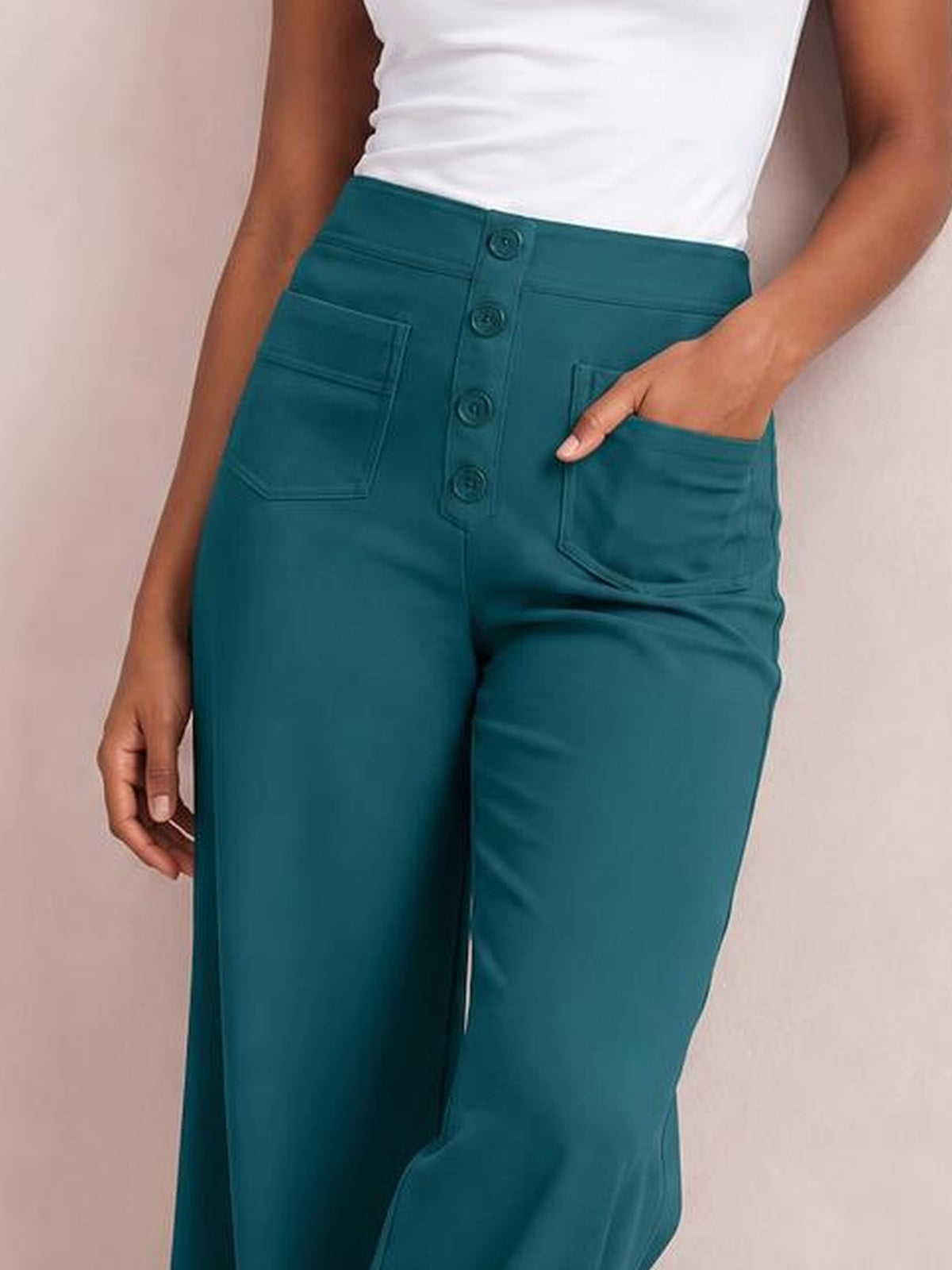 A model wears teal high-waist wide-leg pants with a button-front fly and patch pockets, resting one hand in a pocket to show the relaxed silhouette.