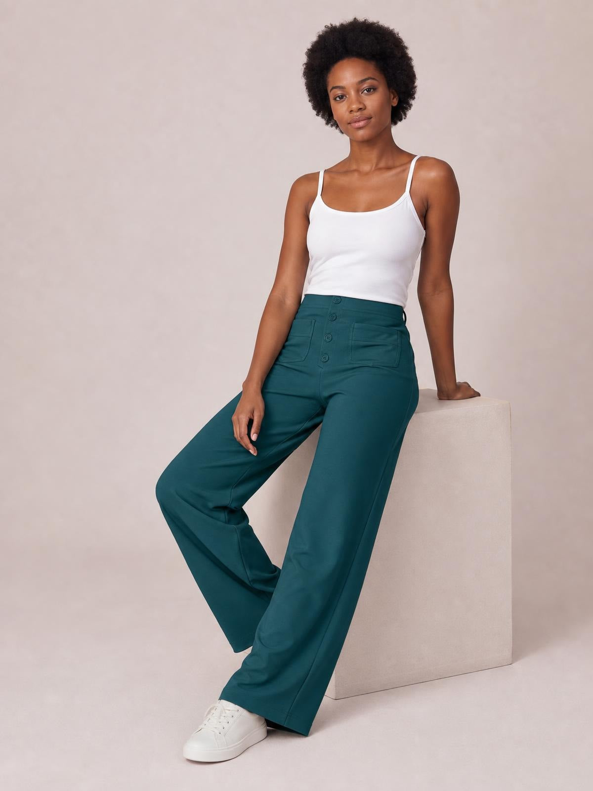 A model poses seated in dark teal high-waist wide-leg pants with a button-front closure and spacious front pockets, showing the relaxed, flowing silhouette.
