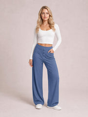 Model wears blue high-waist wide-leg pants with front pockets, button-fly construction, and a relaxed full-length silhouette, posing with one hand in a pocket.