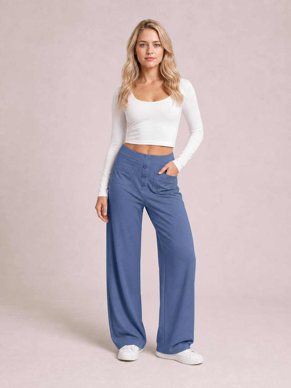 Model wears blue high-waist wide-leg pants with front pockets, button-fly construction, and a relaxed full-length silhouette, posing with one hand in a pocket.
