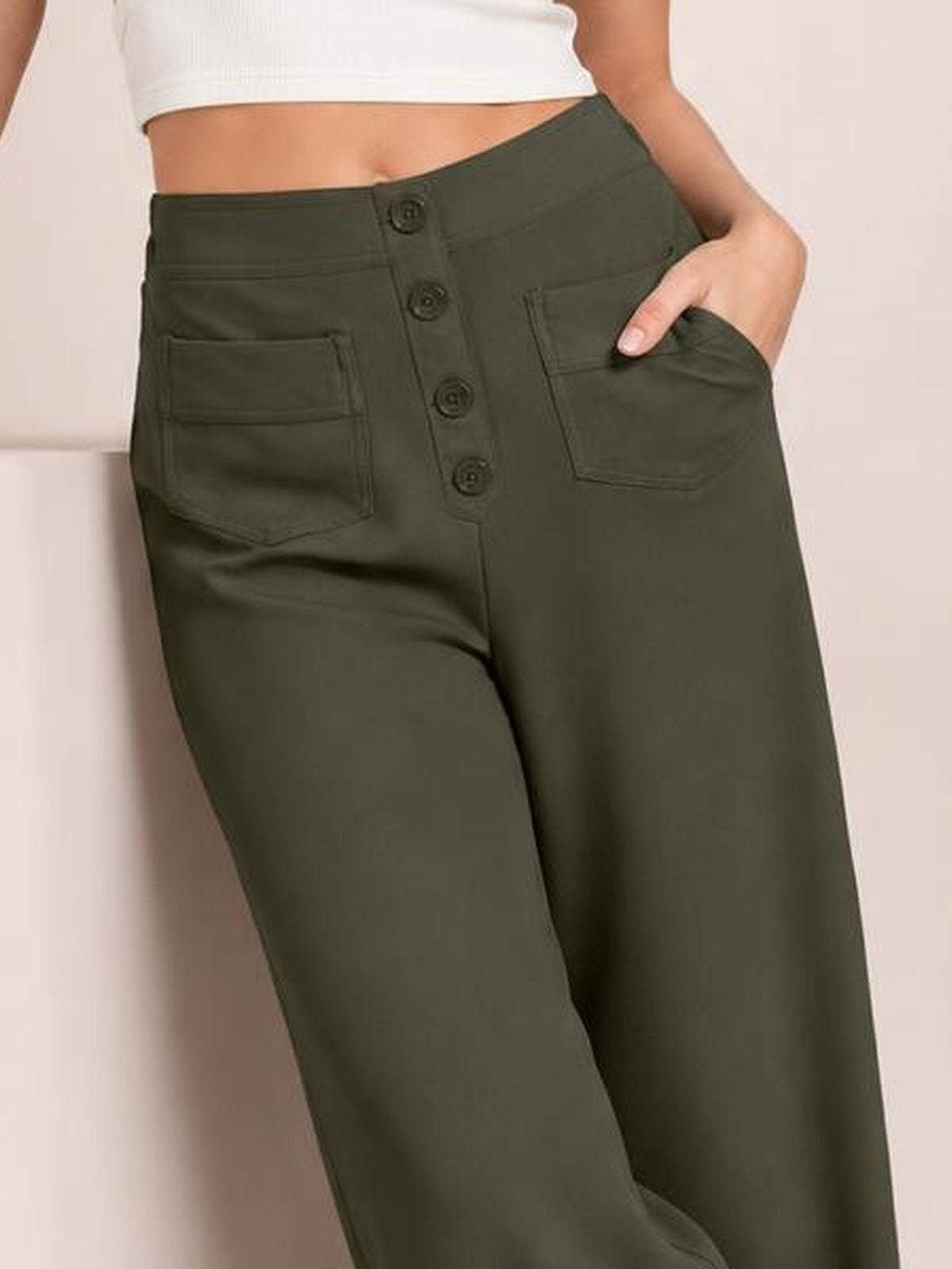 Model wearing dark olive high-waist wide-leg pants with a button fly and two front patch pockets, posing with one hand tucked into a pocket.