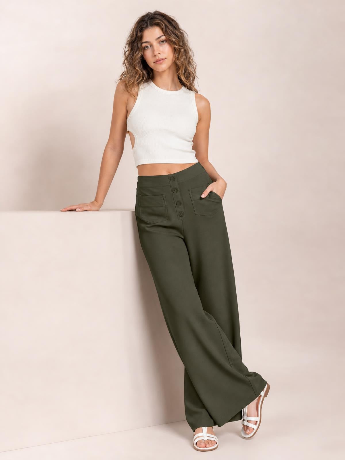Model wears olive green high waist wide leg pants with front button fastening and pockets, posing with one hand in a pocket and legs crossed.
