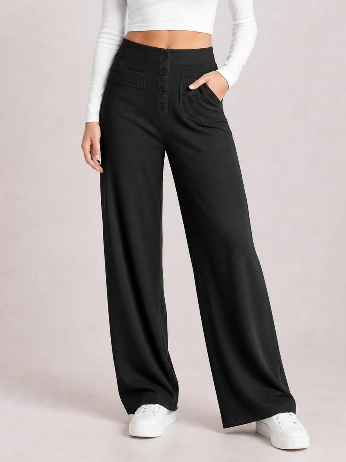 Model showcases black high-waist wide-leg pants with front button closure, stitched pockets, and a flowing full-length silhouette, standing with one hand in a pocket.