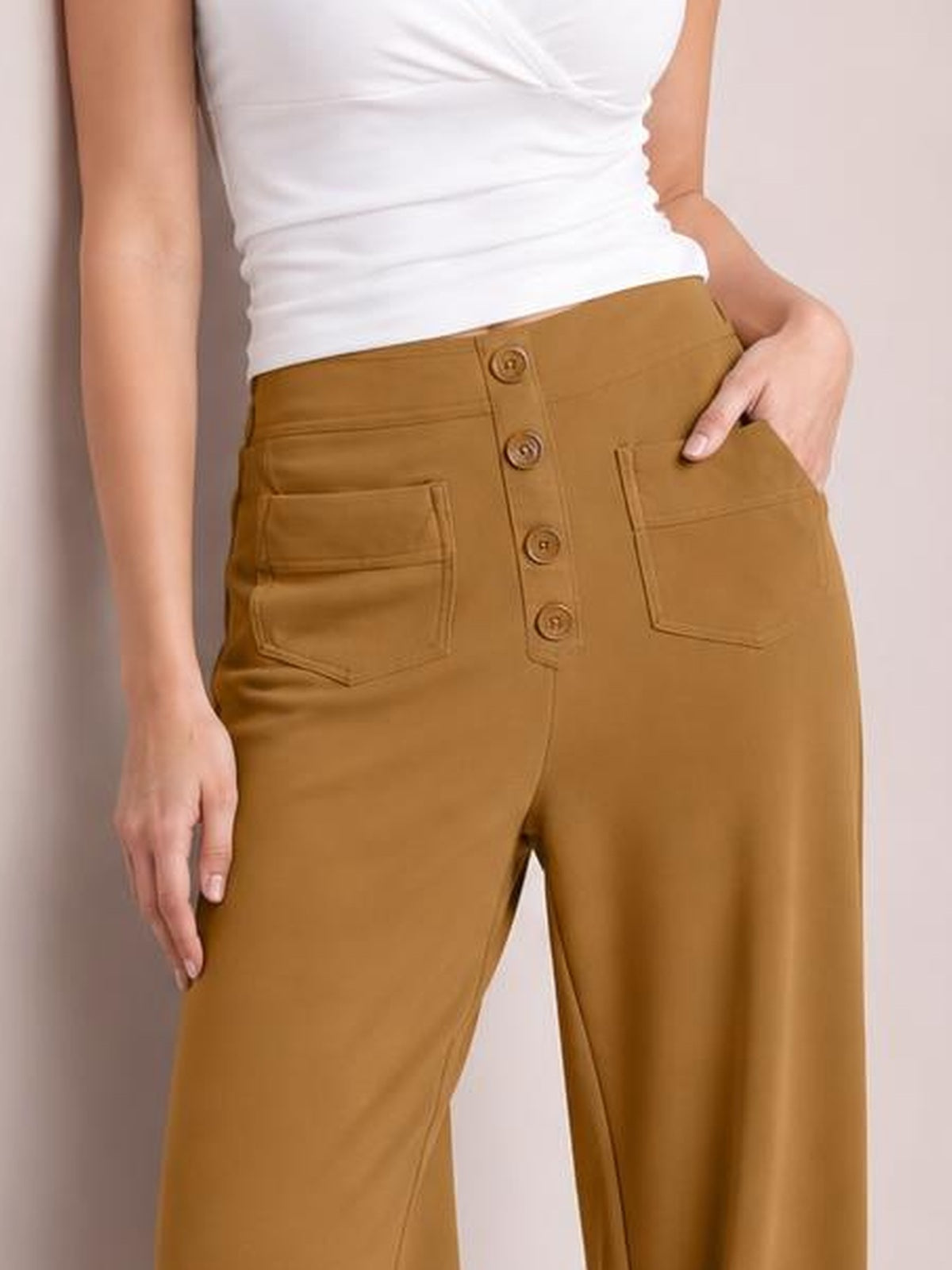 A model wears mustard brown high waist wide leg pants with front button closure and large patch pockets, standing with one hand in a pocket.