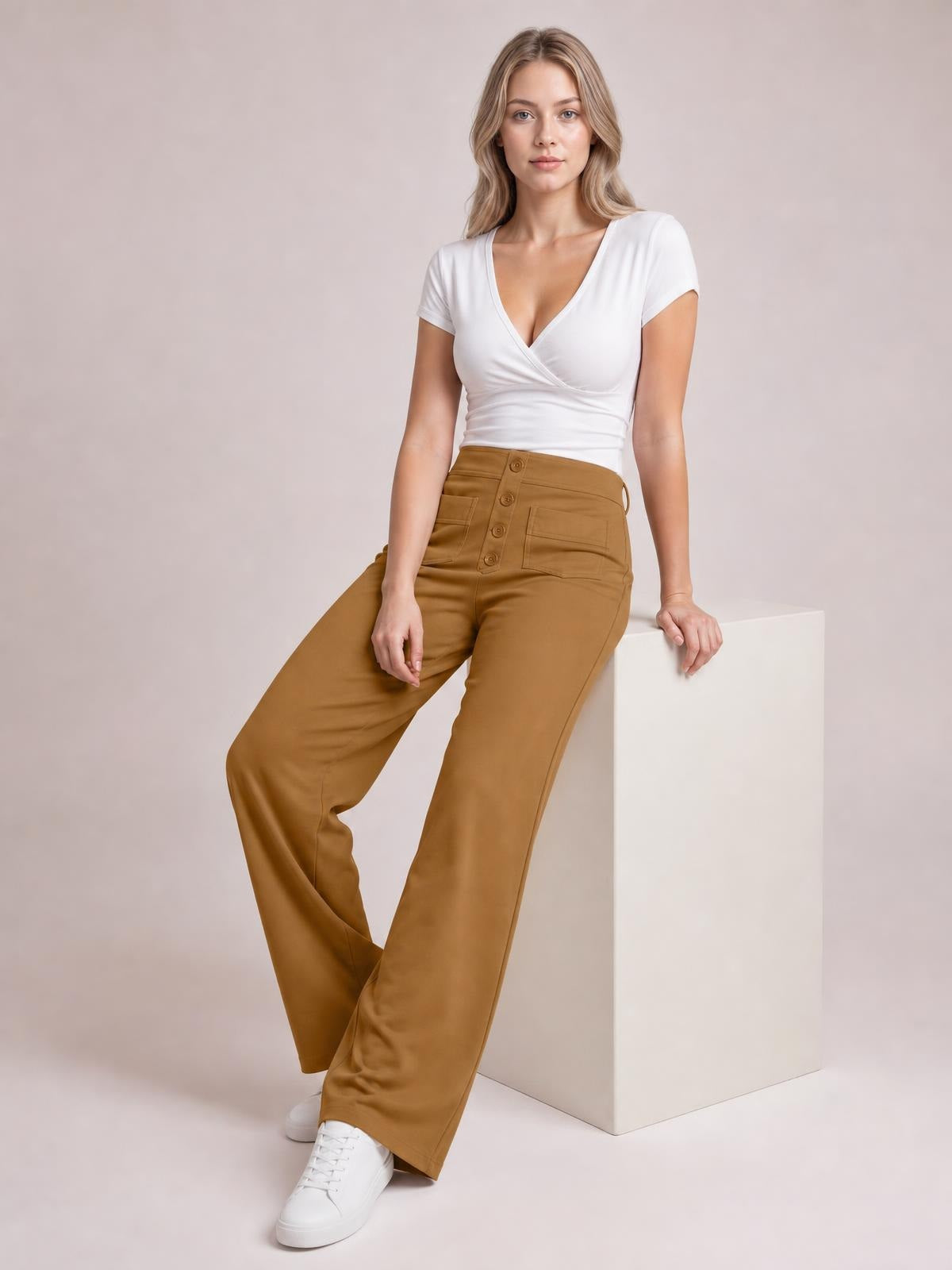 Model wears caramel high waist wide leg pants with front pockets and button closure, posing with one leg crossed while leaning against a cube.
