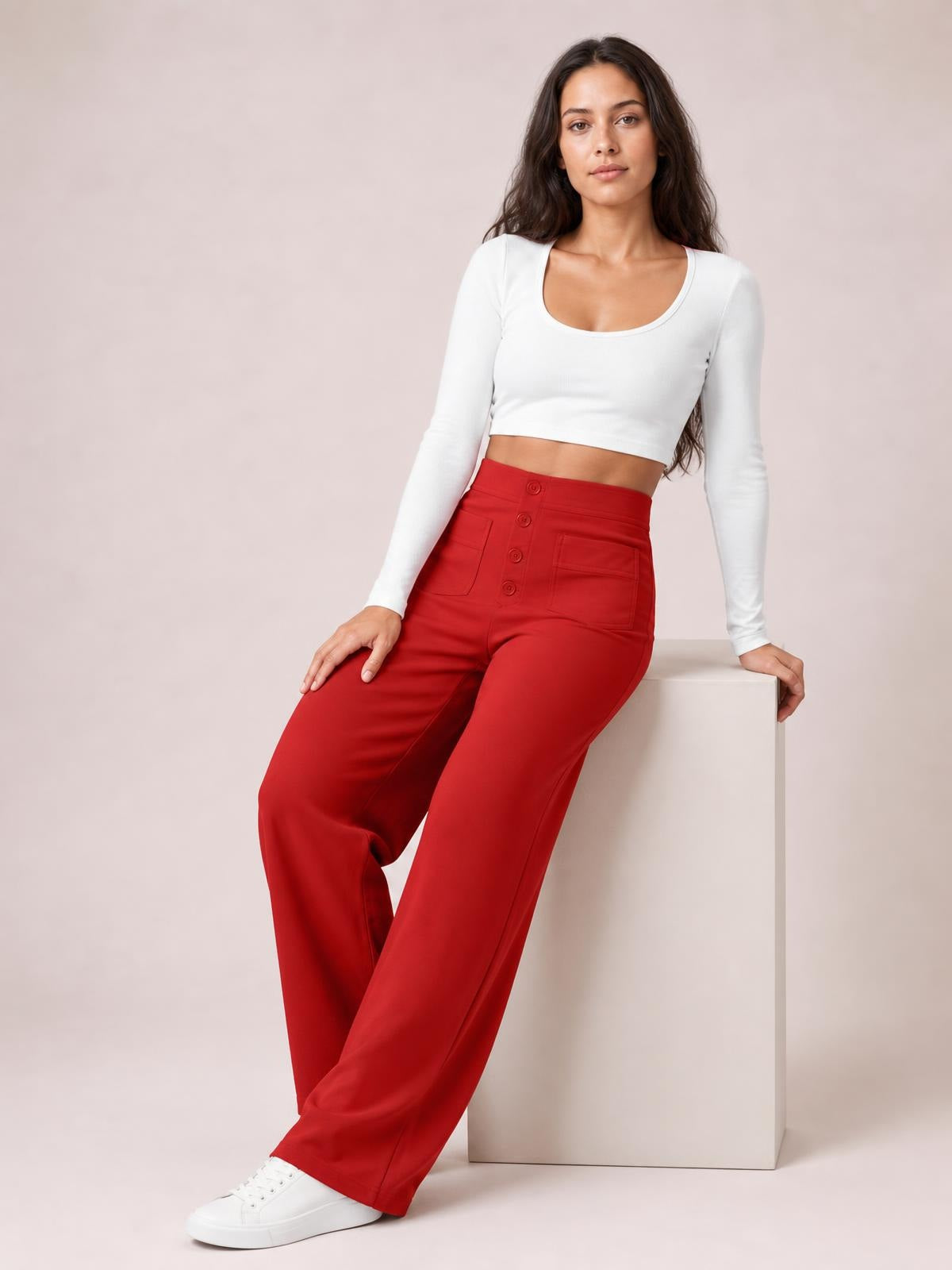 Model seated in red high waist wide leg pants with front pockets and button fly, styled with a white fitted long-sleeve top and sneakers.