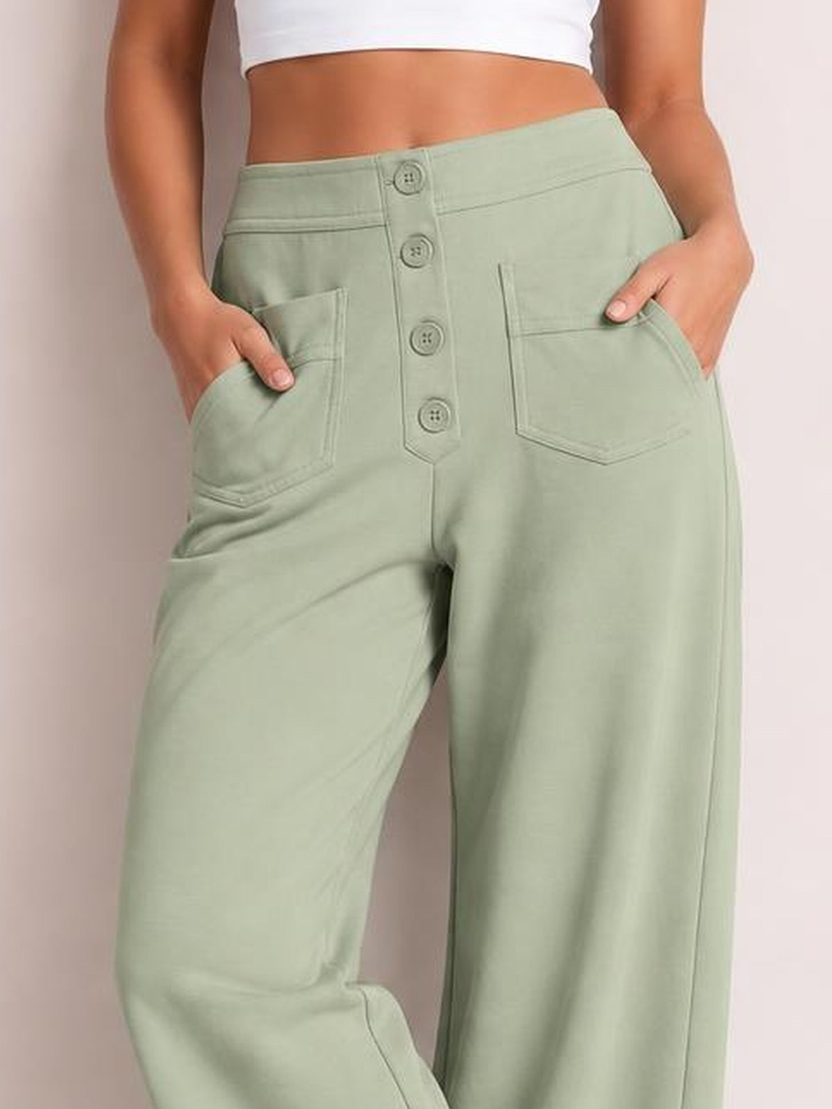 A model wears light sage green high waist wide leg pants with front pockets, a button fly, belt loops, and a relaxed, structured silhouette.