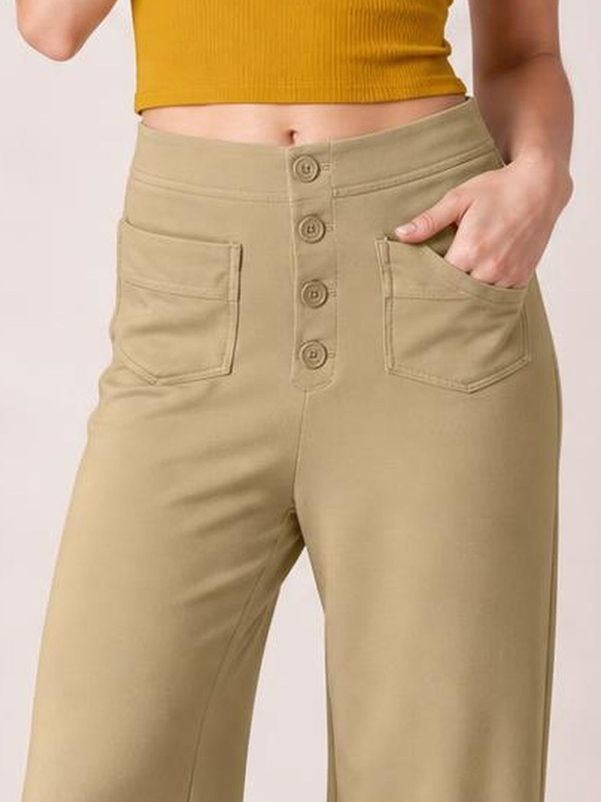 A model wears tan high-waist wide-leg pants with a button fly and front pockets, resting one hand in a pocket to show the relaxed silhouette.